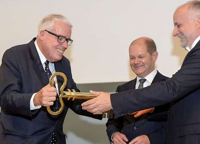 Klaus-Michael Kühne erhält einen großen, goldenen Schlüssel von einem anderen Mann, mit Olaf Scholz im Hintergrund.