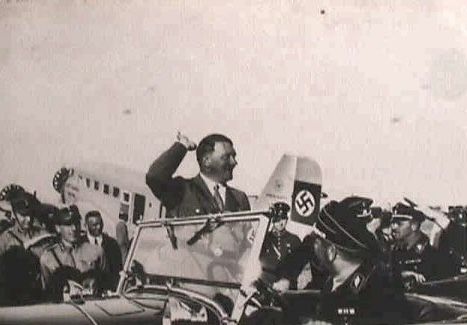 Adolf Hitler winkt in einem offenen Wagen, im Hintergrund ein Flugzeug mit Hakenkreuz.