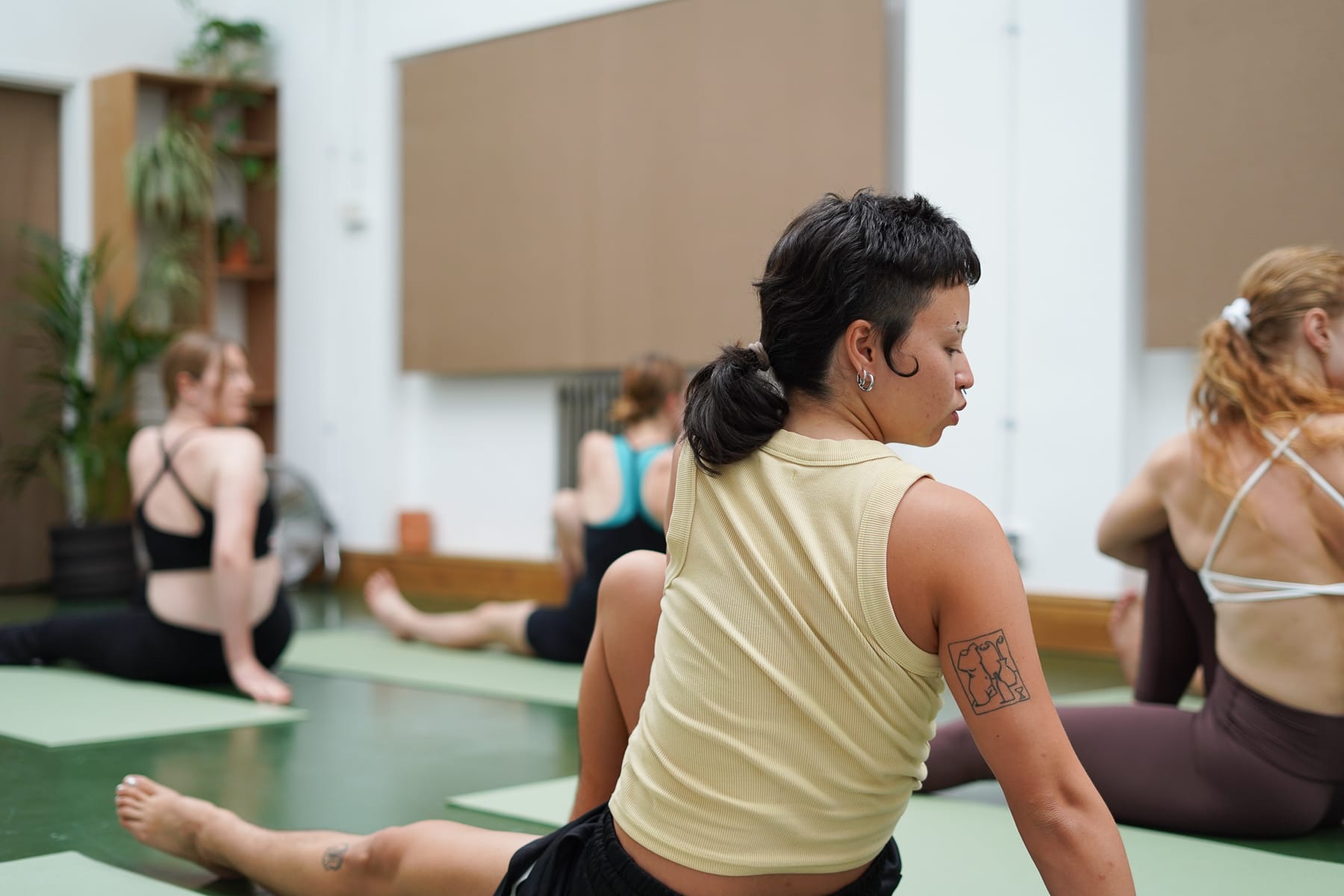 People in workout clothes stretching on yoga mats in a bright studio with plants, a woman with a short dark ponytail in the foreground