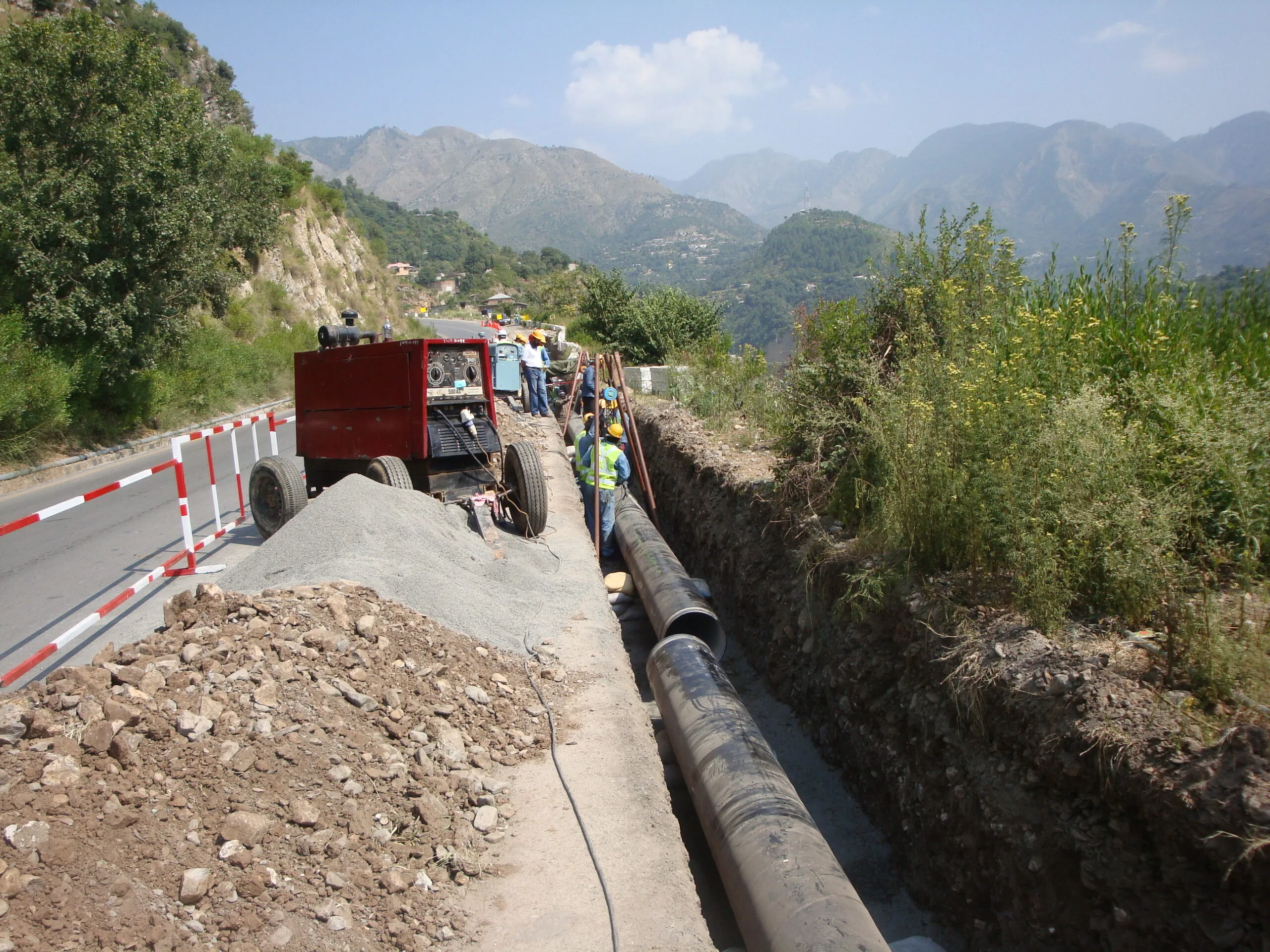Welded steel water main being laid in a rock trench on hilly terrain, welding rig and crew in high-visibility vests