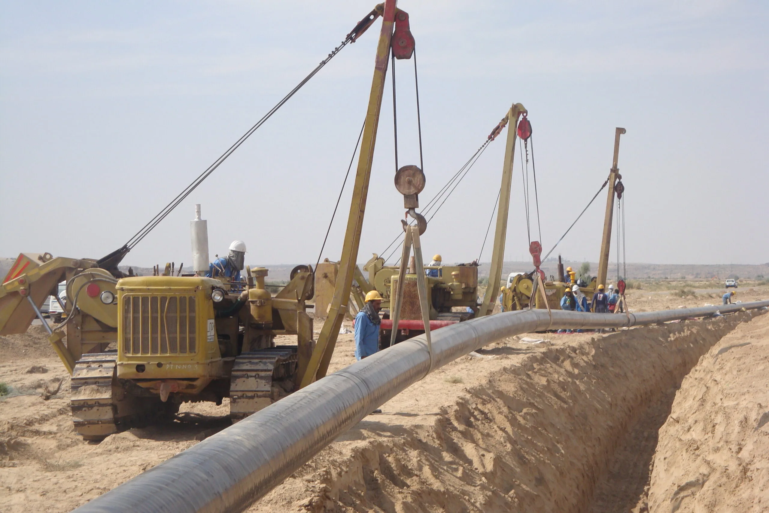 Three side-boom tractors lowering a coated cross-country pipeline into a desert trench, Petrocon crew in hard hats alongside
