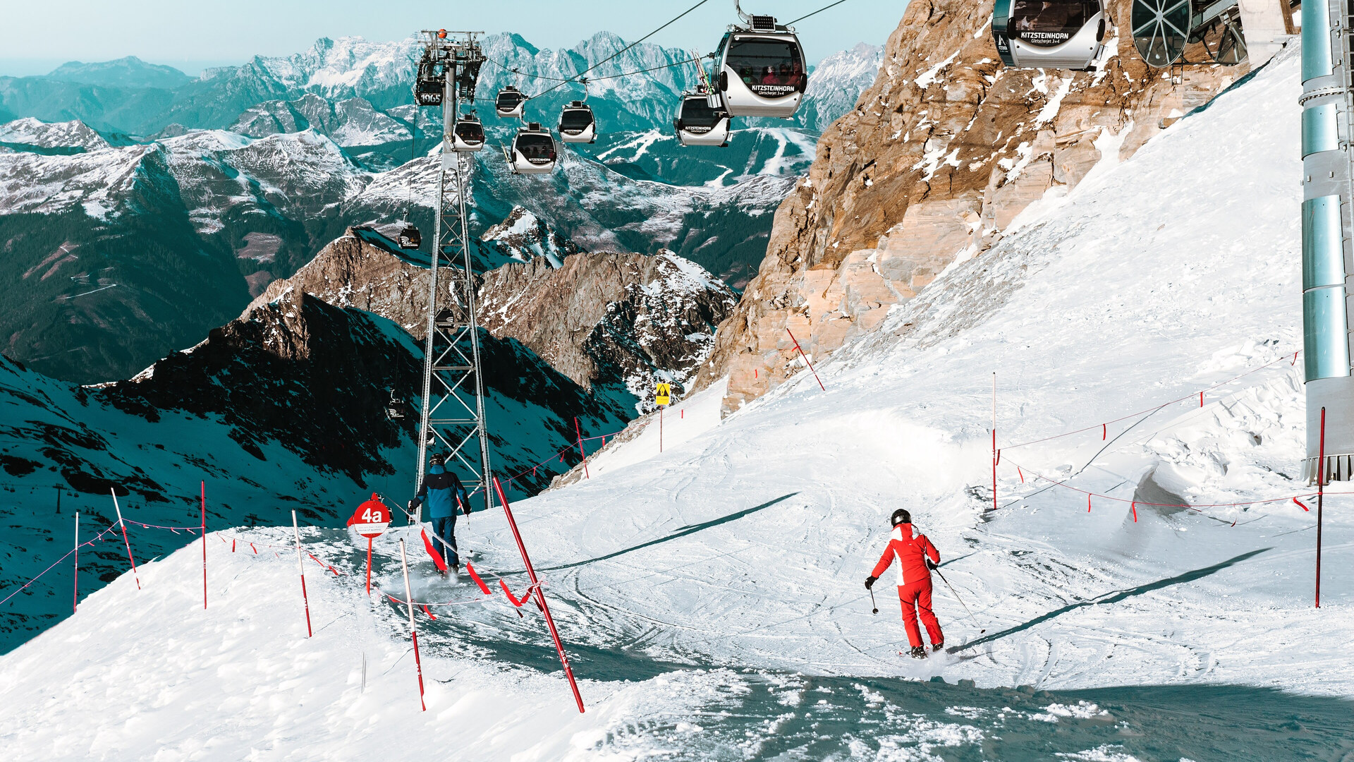 Een skiër in het rood op een geprepareerde gletsjerpiste onder een gondelbaan op het Kitzsteinhorn, met besneeuwde toppen op de achtergrond