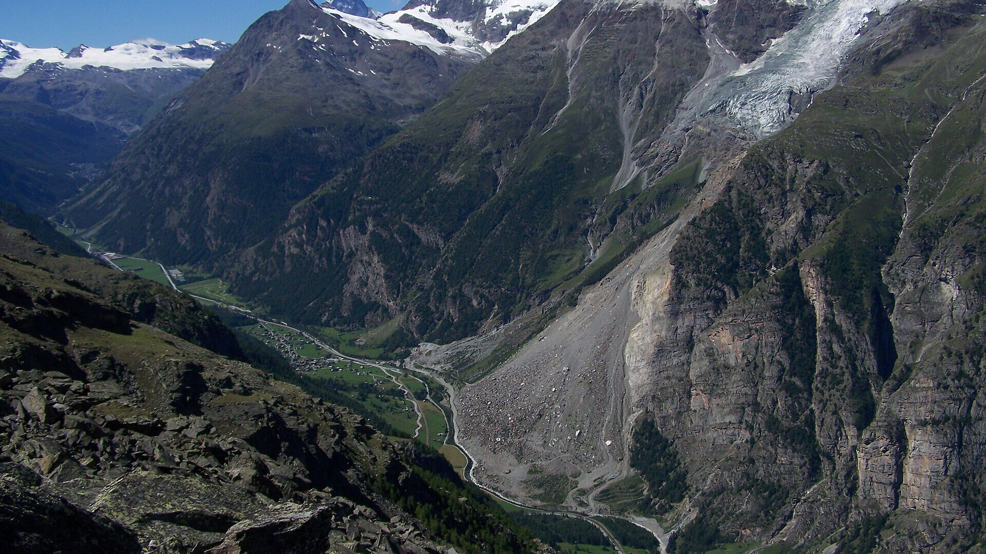 Das Mattertal bei Randa im Sommer, mit dem Dorf auf dem Talboden, dem hellen Schuttkegel des Bergsturzes von 1991 am Westhang und schneebedeckten Gipfeln im Hintergrund