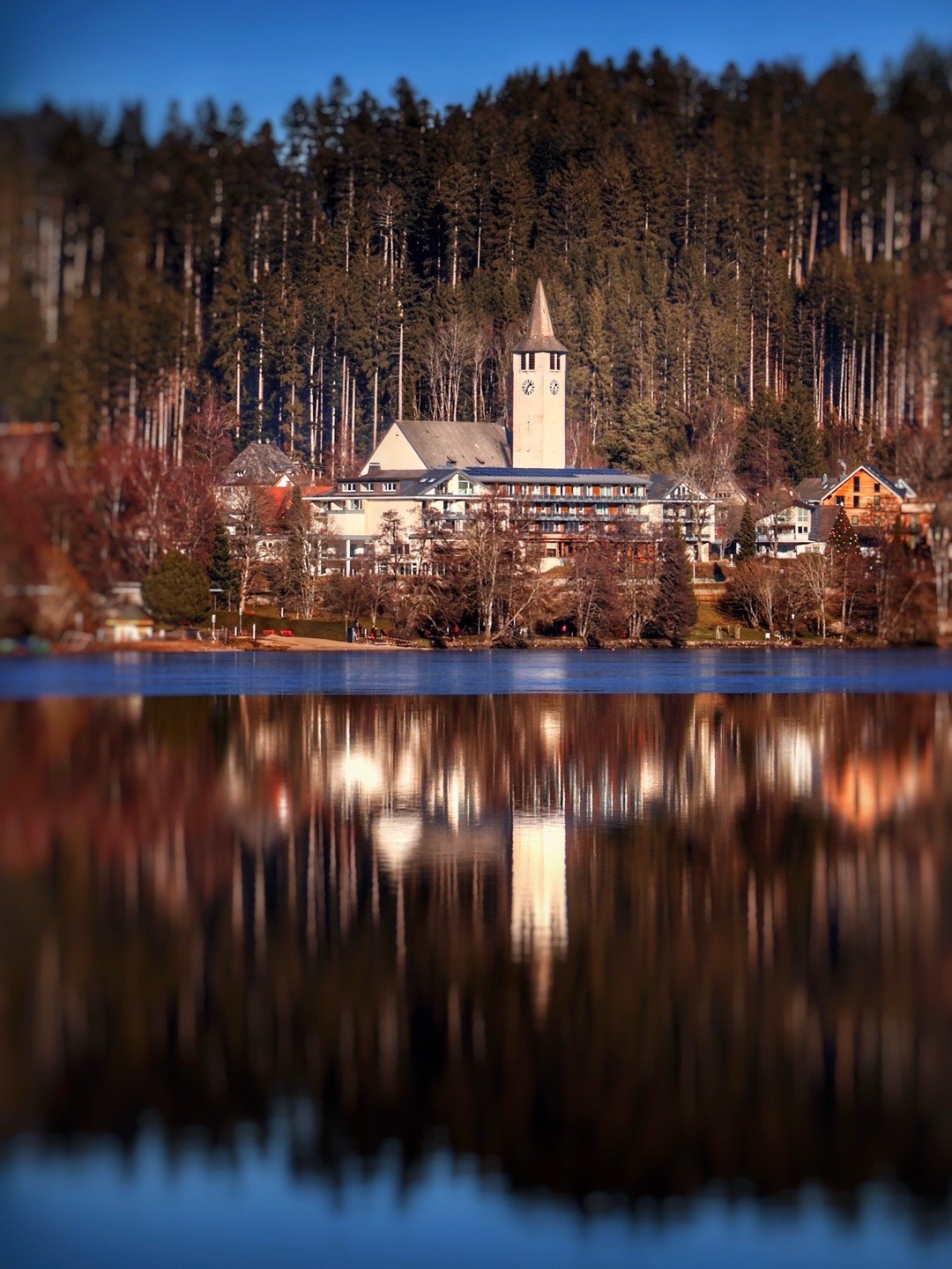 Kirchturm vor Nadelwald mit Spiegelung im Titisee