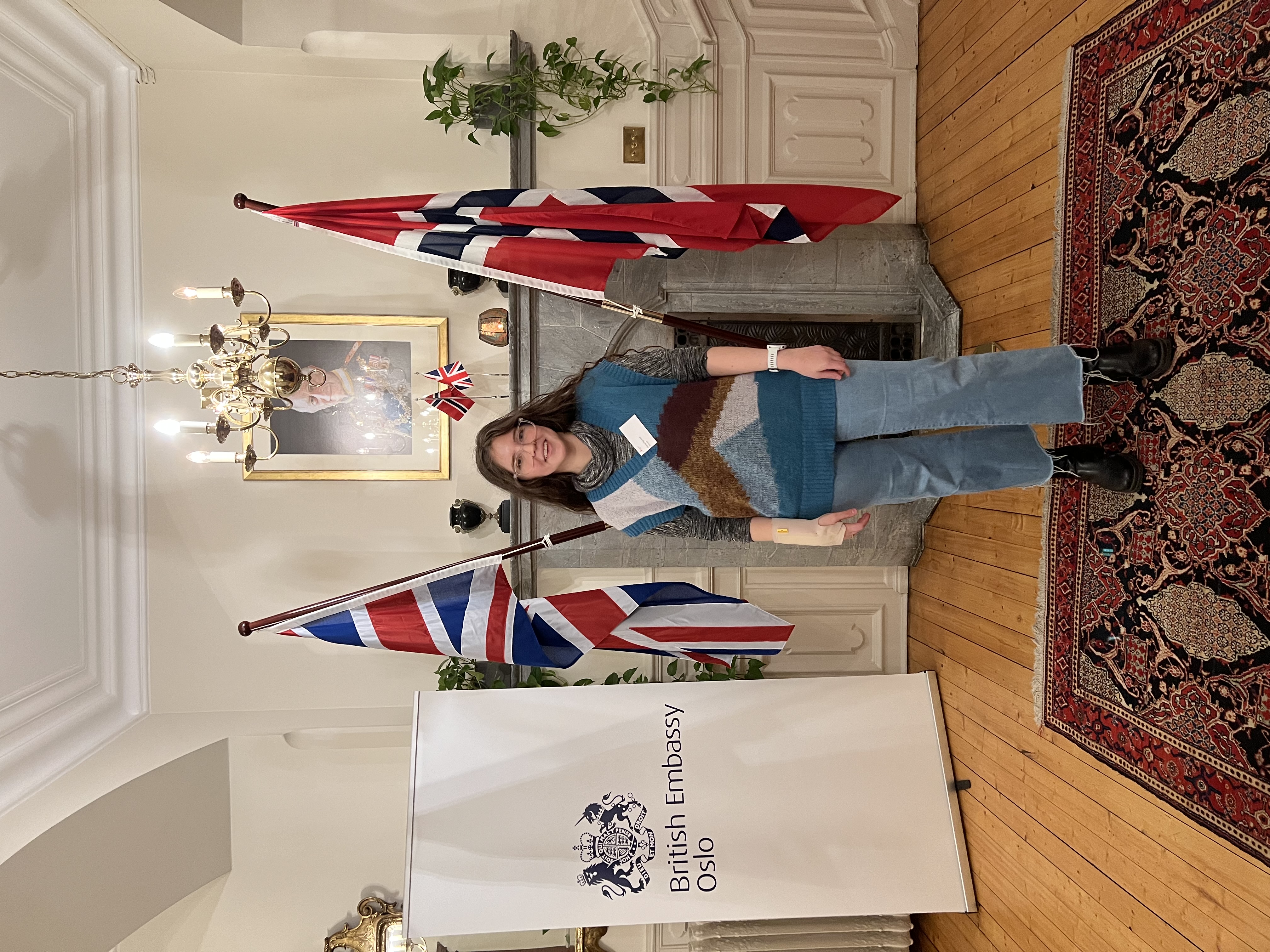 Maria Sigstad, CEO of Plovm, standing in an elegant reception room at the British Embassy Oslo during Oslo Innovation Week. She's wearing a colorful striped sweater in teal, brown and cream tones with blue jeans, positioned between the British Union Jack flag and Norwegian flag. Behind her is a formal fireplace with a crystal chandelier above and a formal portrait on the wall. A 'British Embassy Oslo' banner stands to the left. She's smiling and wearing a name tag, standing on an ornate Persian rug with wooden floors.