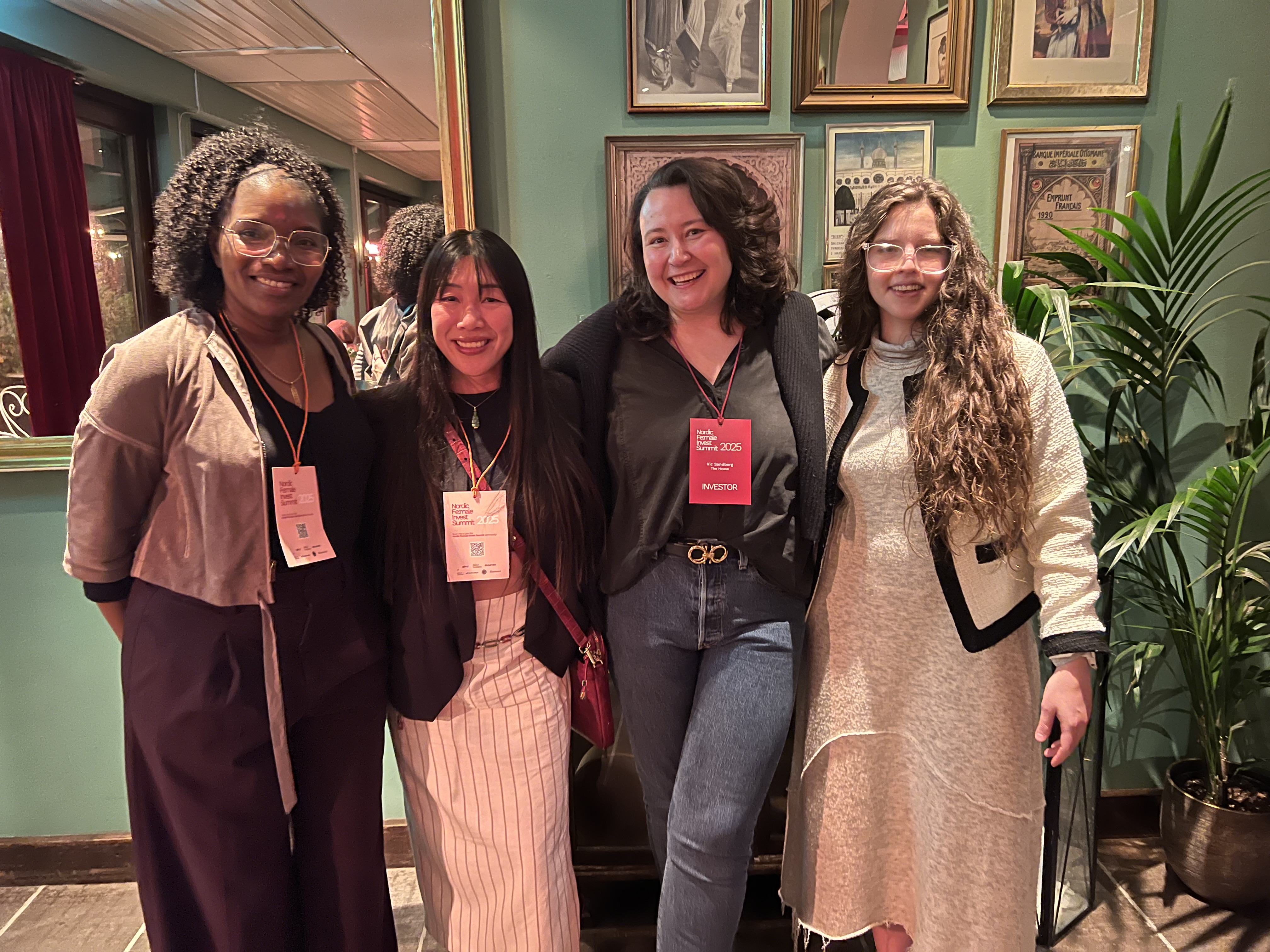Four women standing together smiling at Nordic Fe:male Invest Summit 2025 at Moriskan's Bistro in Malmö. From left to right: Jacqueline Nuwame wearing glasses, beige top and brown vest with event lanyard; a woman in black blazer with white pinstripe skirt and coral event lanyard; Victoria Sandberg in black outfit with red investor badge; and Maria Jimenez Sigstad in cream colored dress with black cardigan. They're standing against a turquoise wall decorated with vintage framed photographs and posters. A large ornate gold-framed mirror reflects the restaurant interior behind them, showing warm lighting and burgundy curtains. A green plant is visible on the right side. The atmosphere is warm and celebratory, capturing a genuine moment of connection between founders at the 10-year anniversary celebration.