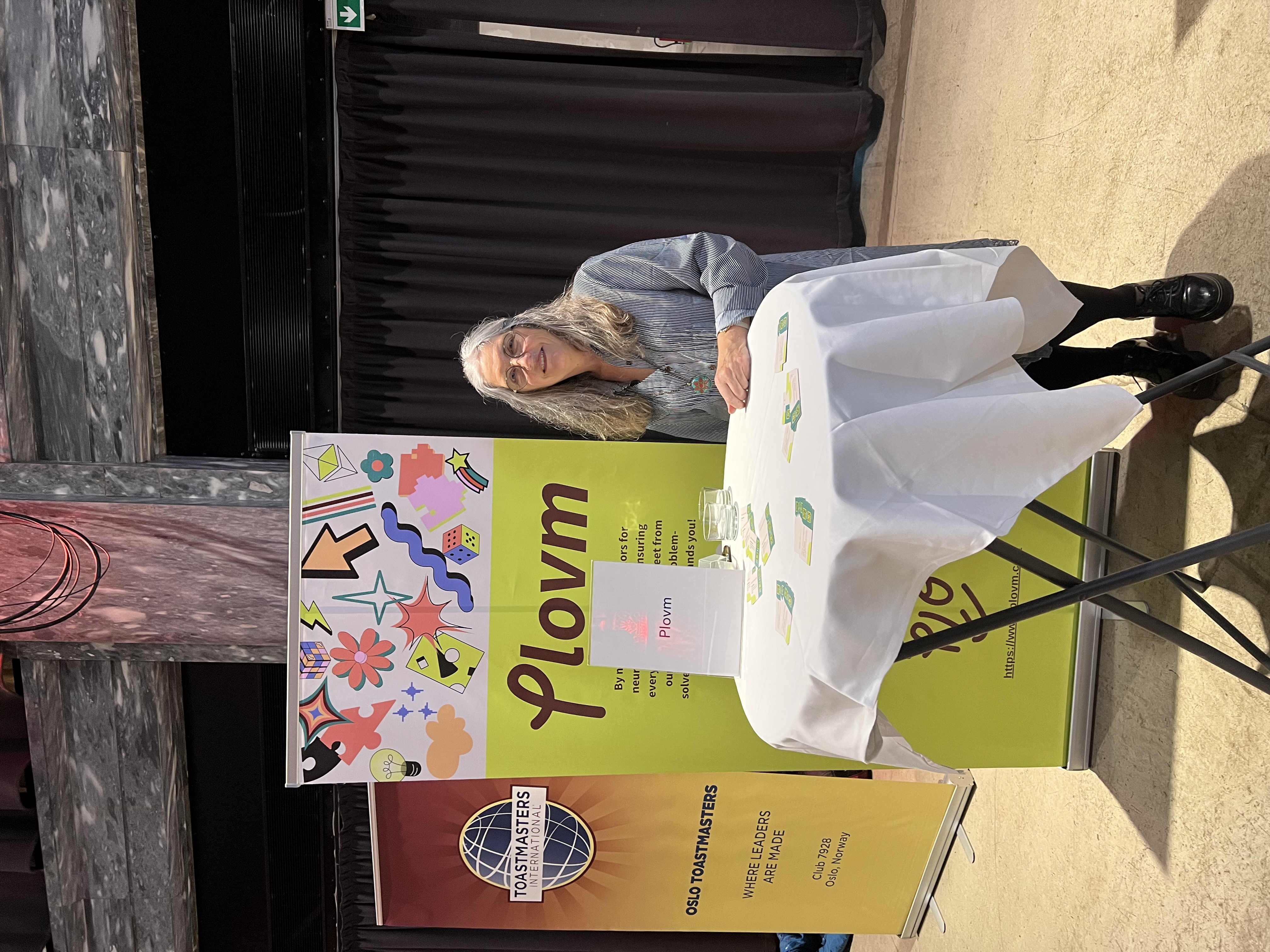 Mariana Sigstad, Plovm's Chief Product Officer, sitting professionally at a round networking table covered with a crisp white tablecloth. She has long, flowing silver-grey hair and wears wire-rimmed glasses with a light blue and white striped button-down collared shirt. Her posture is confident and welcoming as she sits with her hands clasped on the table. Behind her stands a vibrant lime-green promotional banner displaying the Plovm logo prominently, surrounded by colorful decorative elements including bright yellow lightning bolts, orange stars, purple geometric shapes, red dice, pink flowers, blue abstract swirls, green triangles, and various other playful design elements. The banner contains text about Plovm's neurodivergent support services, though partially visible. To her left, a brown and orange Toastmasters International banner displays the organization's distinctive blue and white globe logo with the text 'OSLO TOASTMASTERS' and 'WHERE LEADERS ARE MADE' along with 'Club 7578 Oslo, Norway.' Several green business cards are scattered across the white tablecloth surface. The setting appears to be inside a conference hall or community center with beige flooring and neutral-colored walls.
