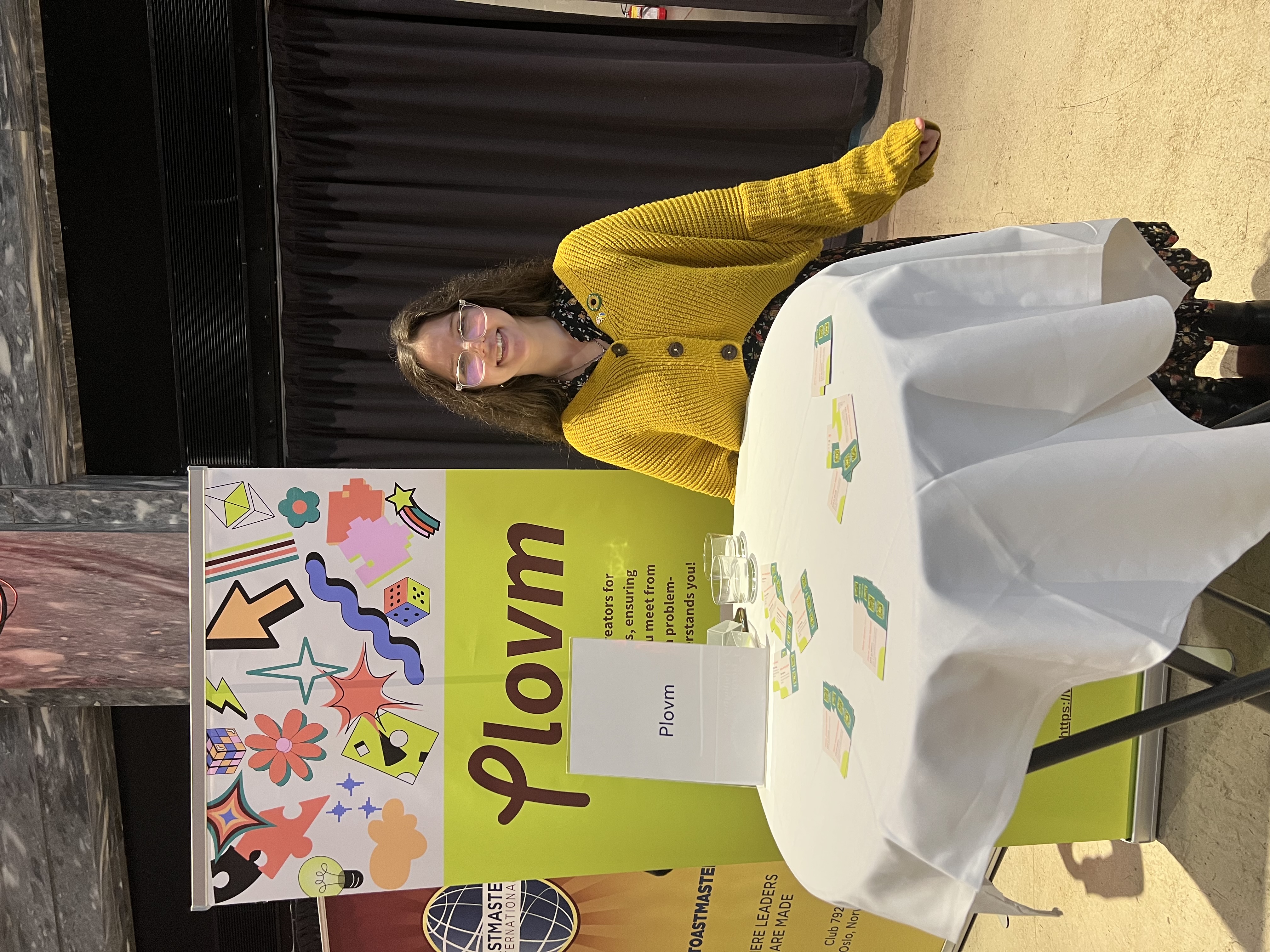 Maria Sigstad, Plovm's Chief Executive Officer, smiling warmly at the same round networking table setup. She wears a bright mustard yellow chunky knit cardigan with large buttons over a dark floral or patterned dress or blouse underneath. Her shoulder-length dark brown hair is styled in soft waves, and she wears clear-framed glasses. Her expression is friendly and approachable as she sits with one hand resting on the table. The same vibrant lime-green Plovm banner serves as her backdrop, featuring the distinctive Plovm logo and an array of colorful decorative elements including stars, lightning bolts, geometric shapes, flowers, and abstract designs in various bright colors like yellow, orange, purple, red, pink, blue, and green. The brown and orange Toastmasters International banner remains visible to the left with its globe logo and text. Multiple green Plovm business cards are scattered across the white tablecloth. The professional networking environment reflects Plovm's mission of making technology accessible and adaptive for diverse users, with both founders demonstrating their commitment to personal, face-to-face connections in the international Oslo community.