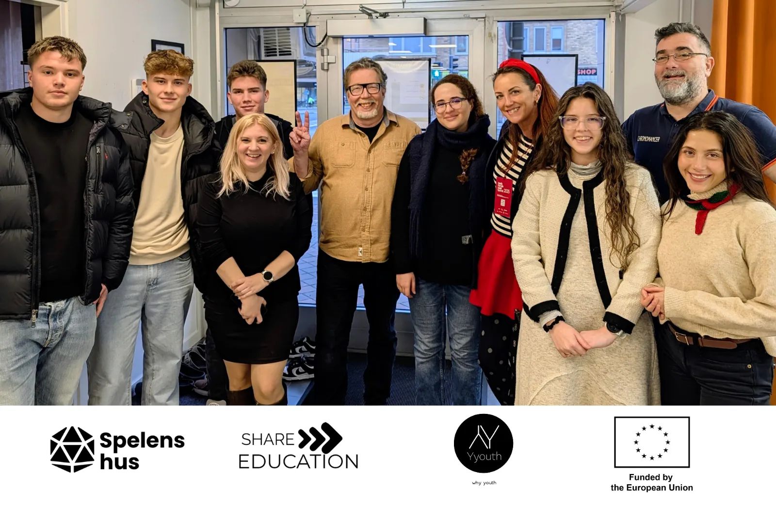 Group photo of international project team at Spelens hus in Malmö, Sweden for BISGIS Erasmus+ project launch. Nine people standing together in modern office space with large windows showing urban buildings in background. Team includes representatives from Sweden (Spelens hus), Norway (Yyouth), and Romania (Share Education). From left: three young men in casual wear, blonde woman in black dress, older man with glasses in brown shirt making peace sign, woman with glasses in dark blazer, woman with long brown hair in striped top and red cardigan, young woman with long brown hair in cream sweater, older man with beard in navy shirt, and young woman with dark hair in cream top. Modern office setting with industrial ceiling, bright natural lighting. Project focuses on game development and social innovation to combat youth isolation. EU and Erasmus+ logos visible at bottom of image.