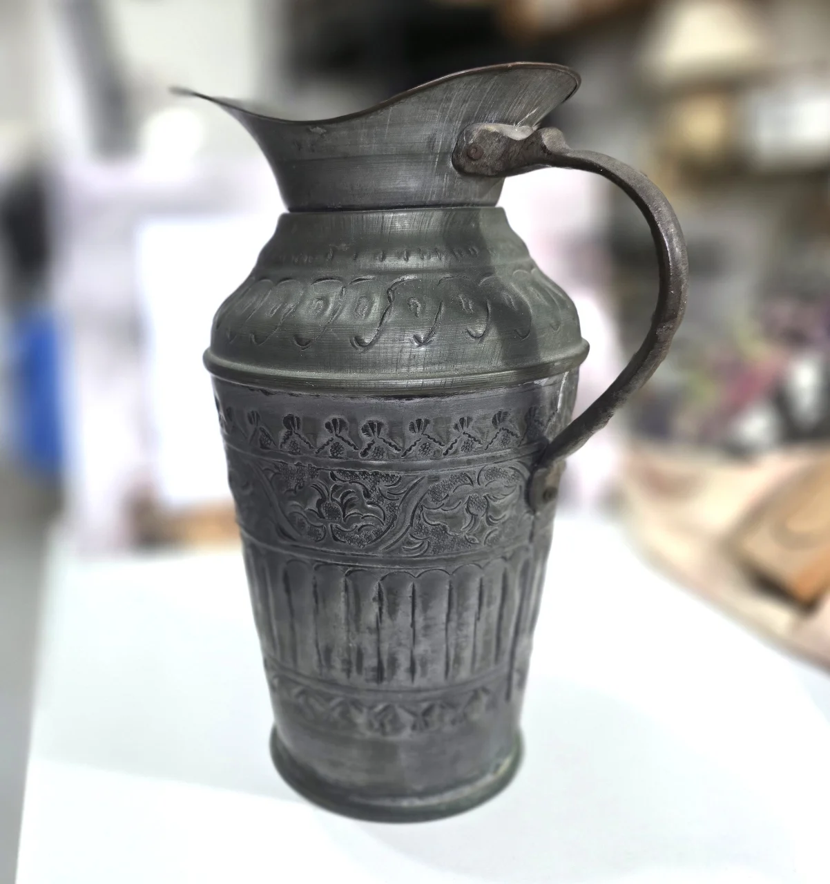 כד נחושת עתיק, מהמאה ה-19, המאופיין בסגנון פרסי או מזרח-תיכוני מסורתי. עשוי נחושת בעבודת יד, (מצופה בדיל) המעניק לו מראה כסוף או אפרפר. הכד מעוטר בדגמים חרוטים (Repoussé) של צמחייה או פרחים, ובעל ידית מעוקלת לנישיאה. גובה 30 סמ