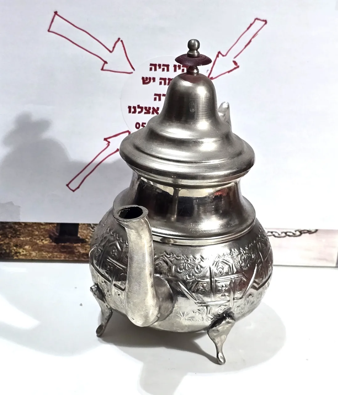 קומקום תה דקורטיבי בסגנון מזרחי ממתכת בגוון כסוף, עומד על שלוש רגליים, עם פיה ומכסה מחובר בציר. עיטור חרוט בתבליט בסגנון מסורתי. מתאים לאספנות, לעיצוב או לשימוש. מצב כללי טוב עם סימני שימוש בהתאם לגיל. מידות: ‎20×24×14 ס”מ