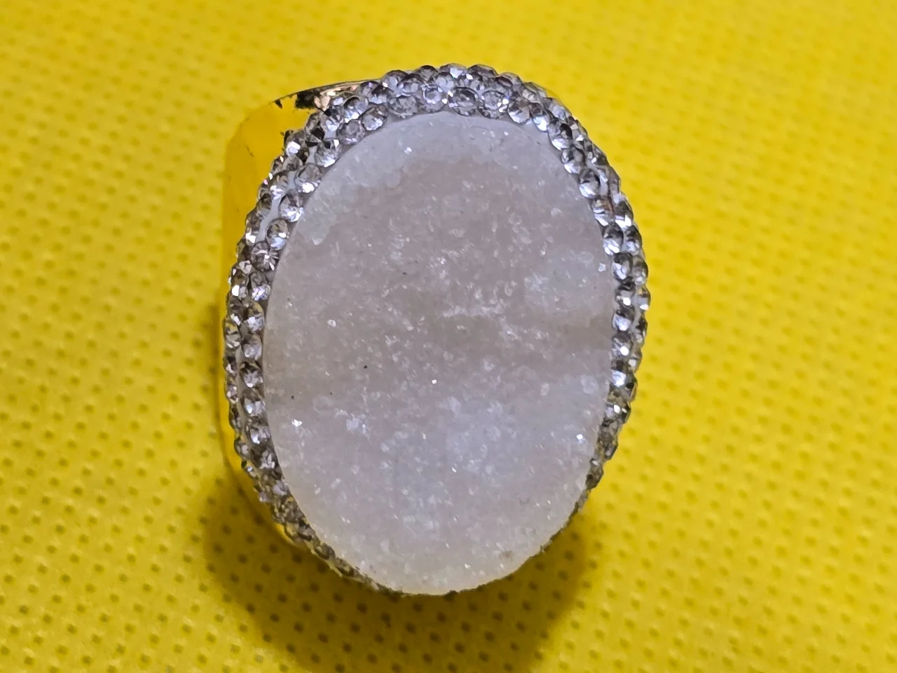 טבעת מדהימה בעבודת יד! משובצת באבן מחוספסת גבישית נוצצת בצבעי לבן קרח. גוף מתכת מוזהב שניתן להתאים לגודל האצבע. מסביב יהלומים סינטטיים נוצצים.