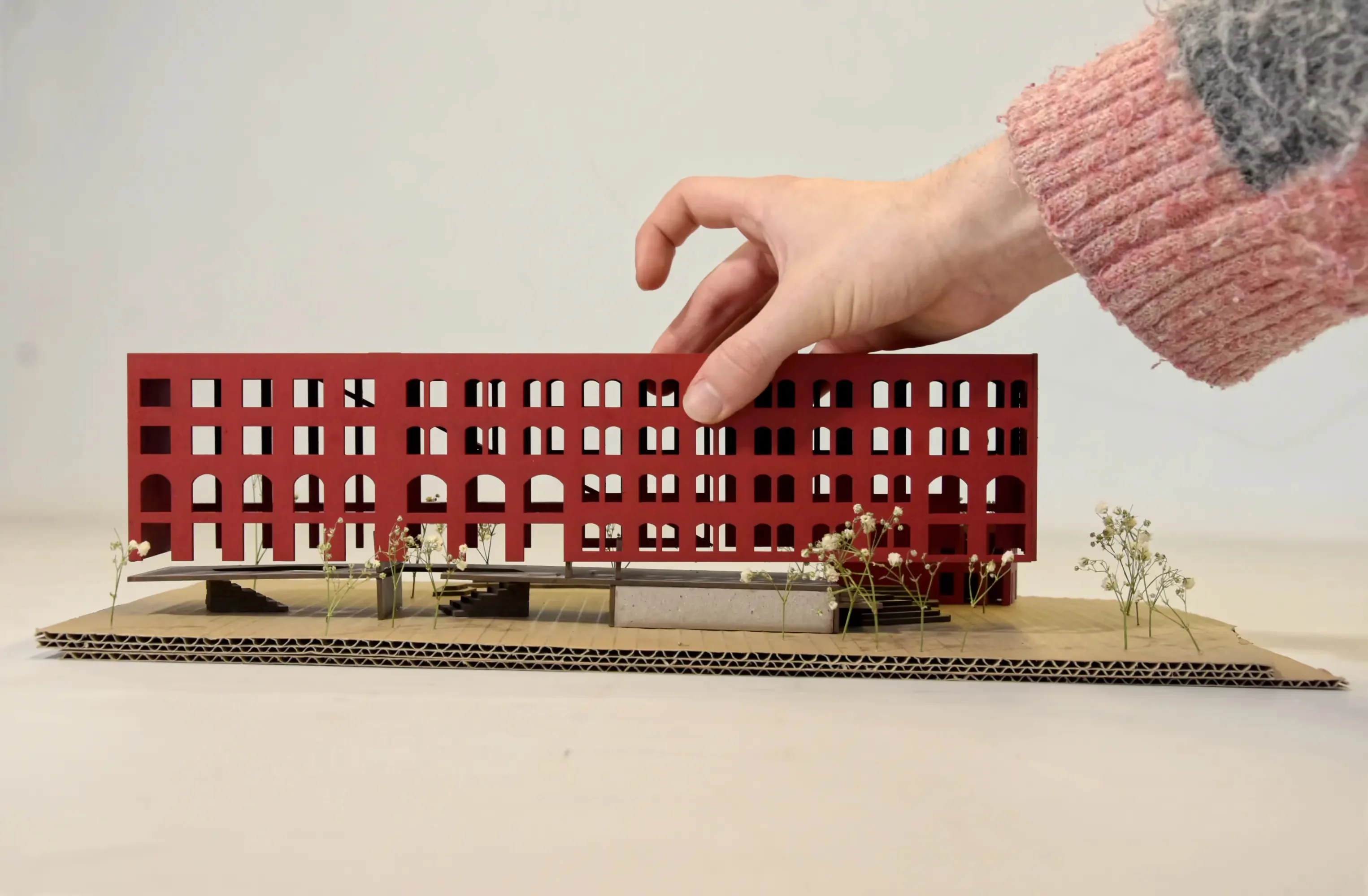 Seitenansicht des Modells: Eine Hand setzt die rote Fabrikfassade präzise über die terrassierte, graue Sockelzone.