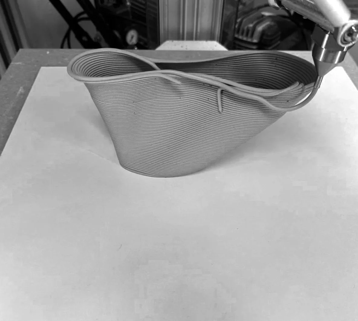 Eine Nahaufnahme des Liquid Deposition Modeling (LDM) Prozesses, bei dem flüssiger Ton schichtweise extrudiert wird. Die Aufnahme dokumentiert eine kritische Phase der Geometrieprüfung, speziell das Verhalten von Überhängen (Cantilevers). Man sieht, wie sich die Tonschichten aufgrund des Eigengewichts und der noch fehlenden Stützung leicht verformen – eine technische Erkenntnis über die Grenzen der Materialviskosität und den notwendigen Neigungswinkel für einen stabilen Druck.