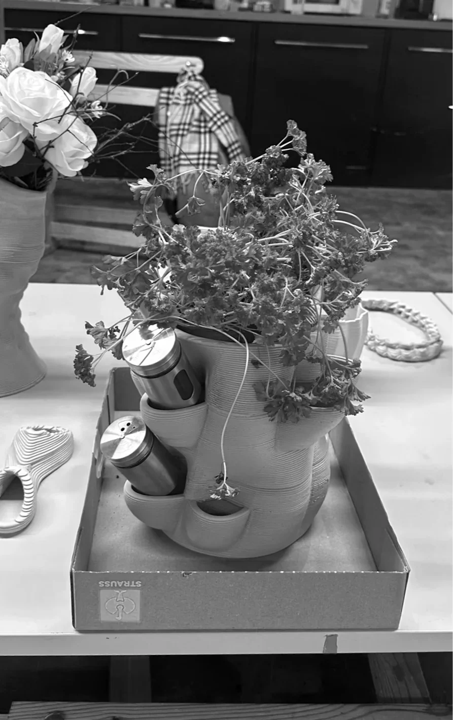 Eine Alltagsszene in einer Küche, die den Spice Cylinder in voller Funktion zeigt. Im oberen Hohlraum wächst frische Petersilie, während in den seitlichen Ton-Balkonen zwei Edelstahl-Gewürzstreuer schräg angedockt sind. Die Visualisierung beweist die Lösung für gängige Frustrationen der Küchenorganisation: maximale Sichtbarkeit und Stabilität bei 360-Grad-Zugänglichkeit. Das Objekt verbindet natürliches Wachstum direkt mit funktionaler Lagerung.