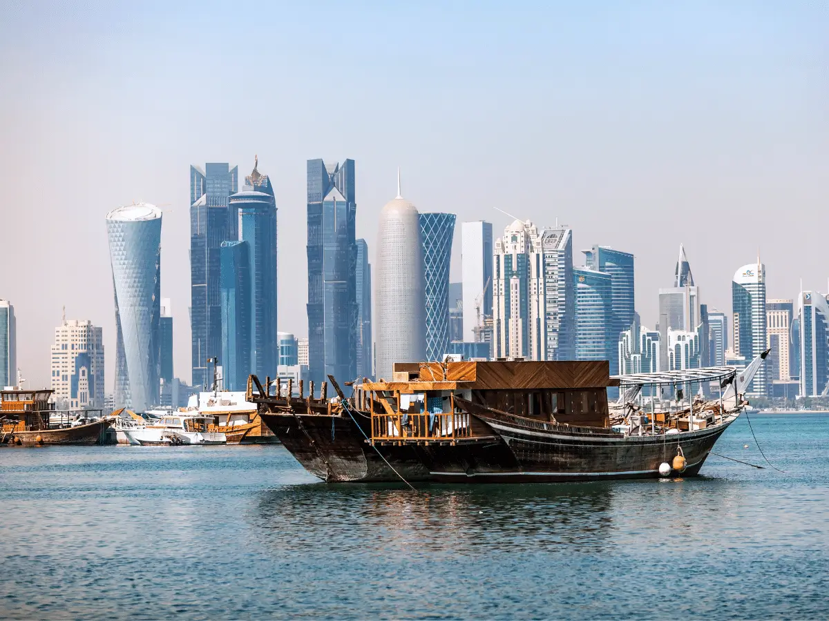Doha skyline view from the Corniche with traditional boats on the waterfront and modern skyscrapers in the background.