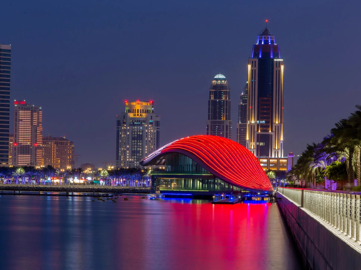 Aerial view of Lusail Promenade along the waterfront with modern walkways, landscaped areas, and nearby architecture, featuring the distinctive shell-inspired Lusail Metro station design.