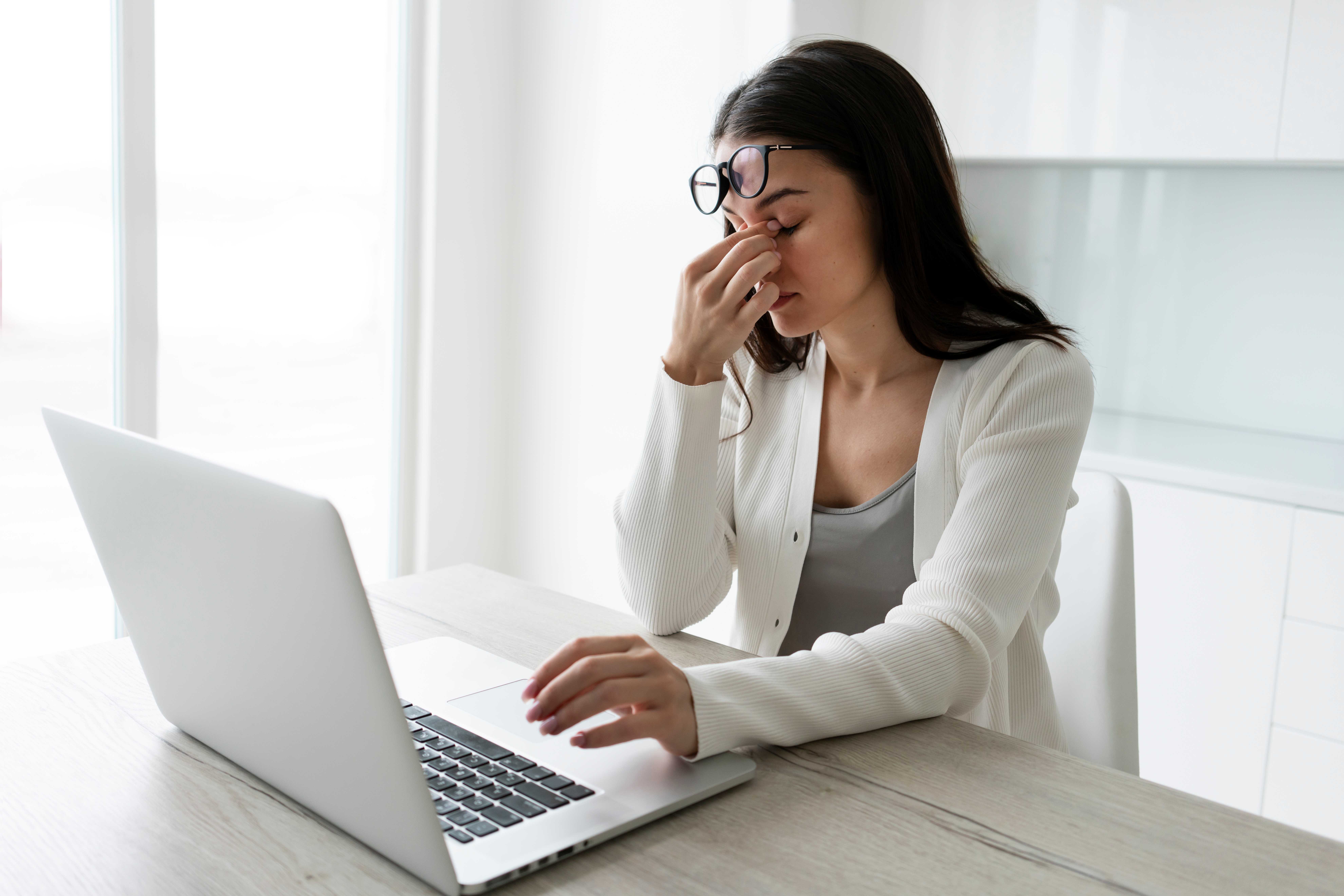 Woman rubbing her eyes while working on a computer with blurred text on screen.