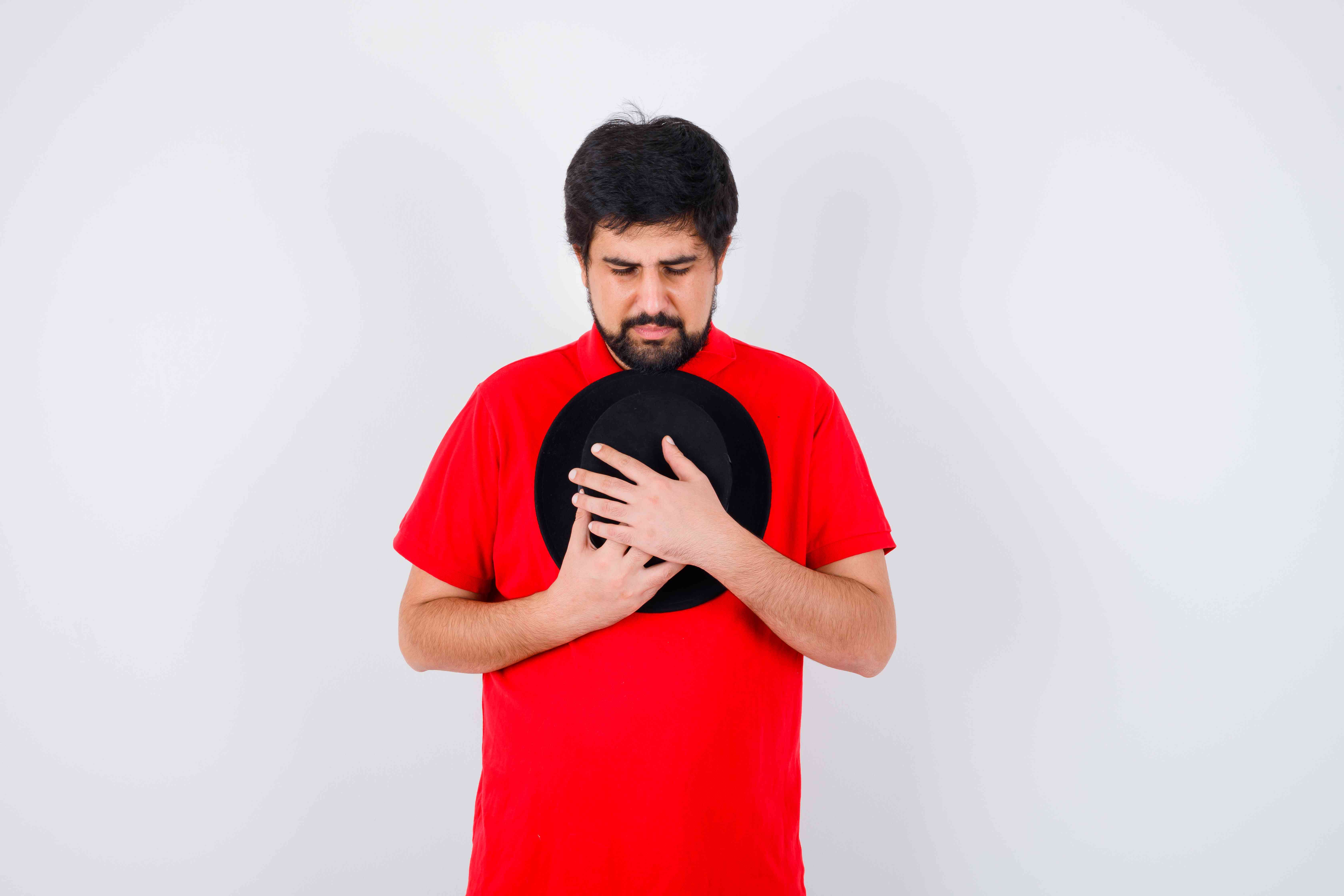 Young man in red shirt holding a hat against his chest with a pained expression.