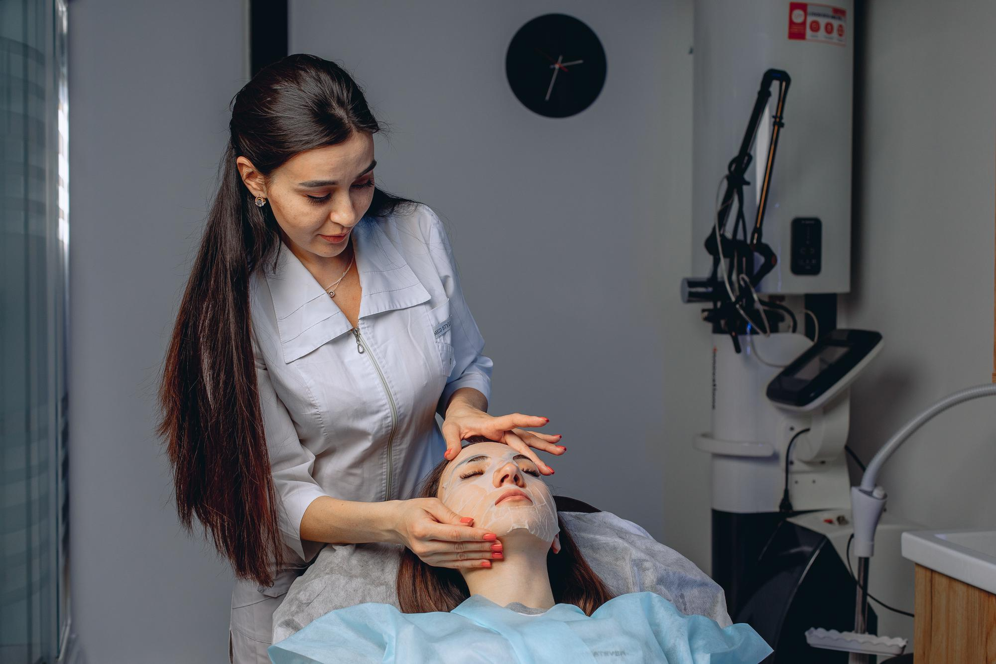 A woman at a dermatologist’s clinic while the doctor examines her facial skin and puts on a face mask.