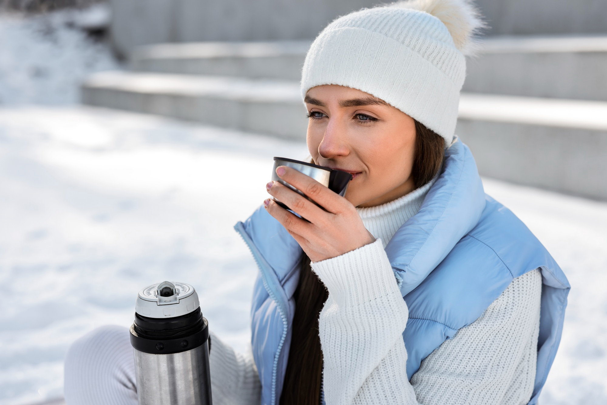 A woman sitting outside in the snow, wearing a winter cap, sweater and jacket, drinking warm water from a thermos.