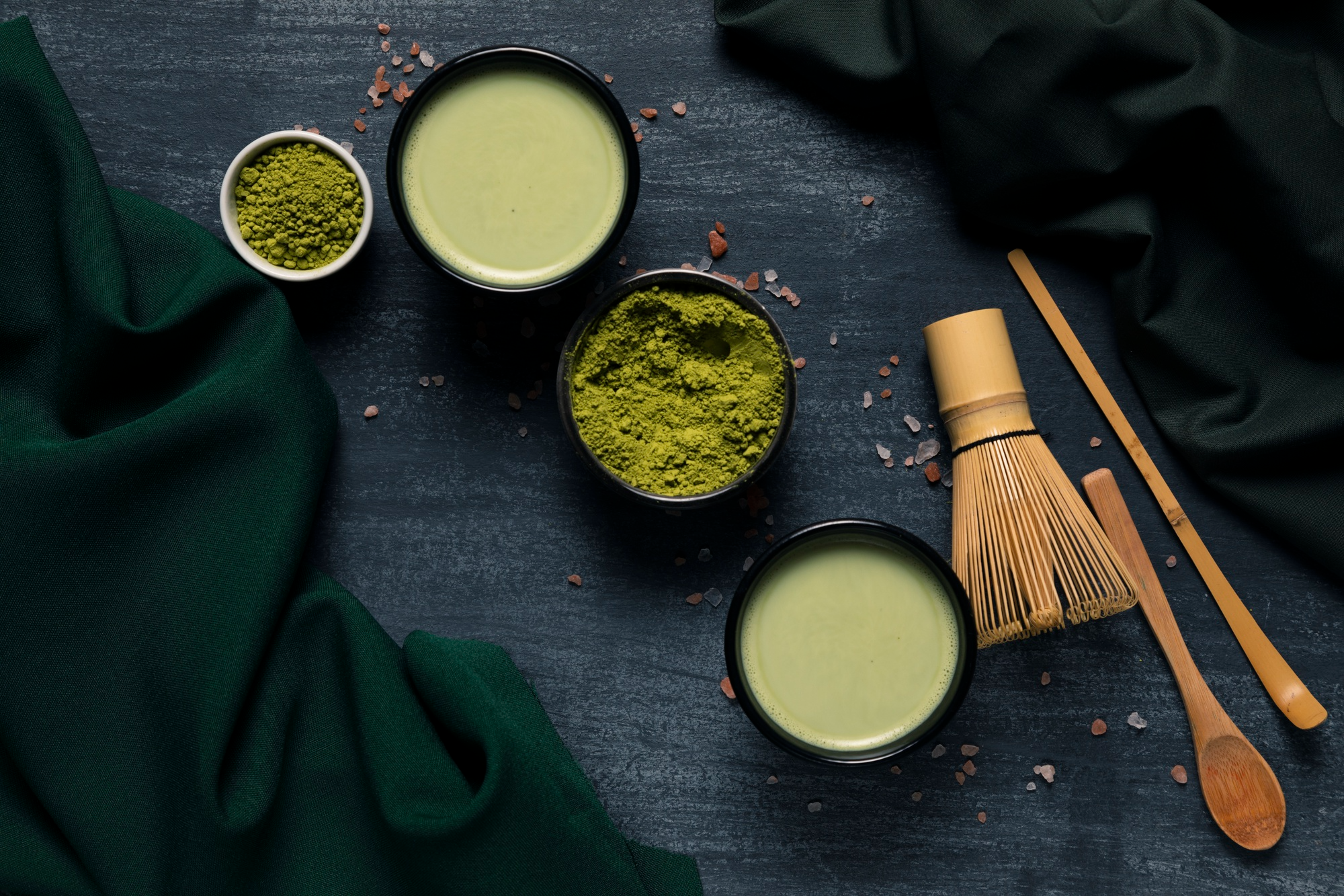 Bright green matcha powder in a bowl with two cups of matcha with a bamboo whisk and a spoon beside it, antioxidant rich superfood for energy and focus.