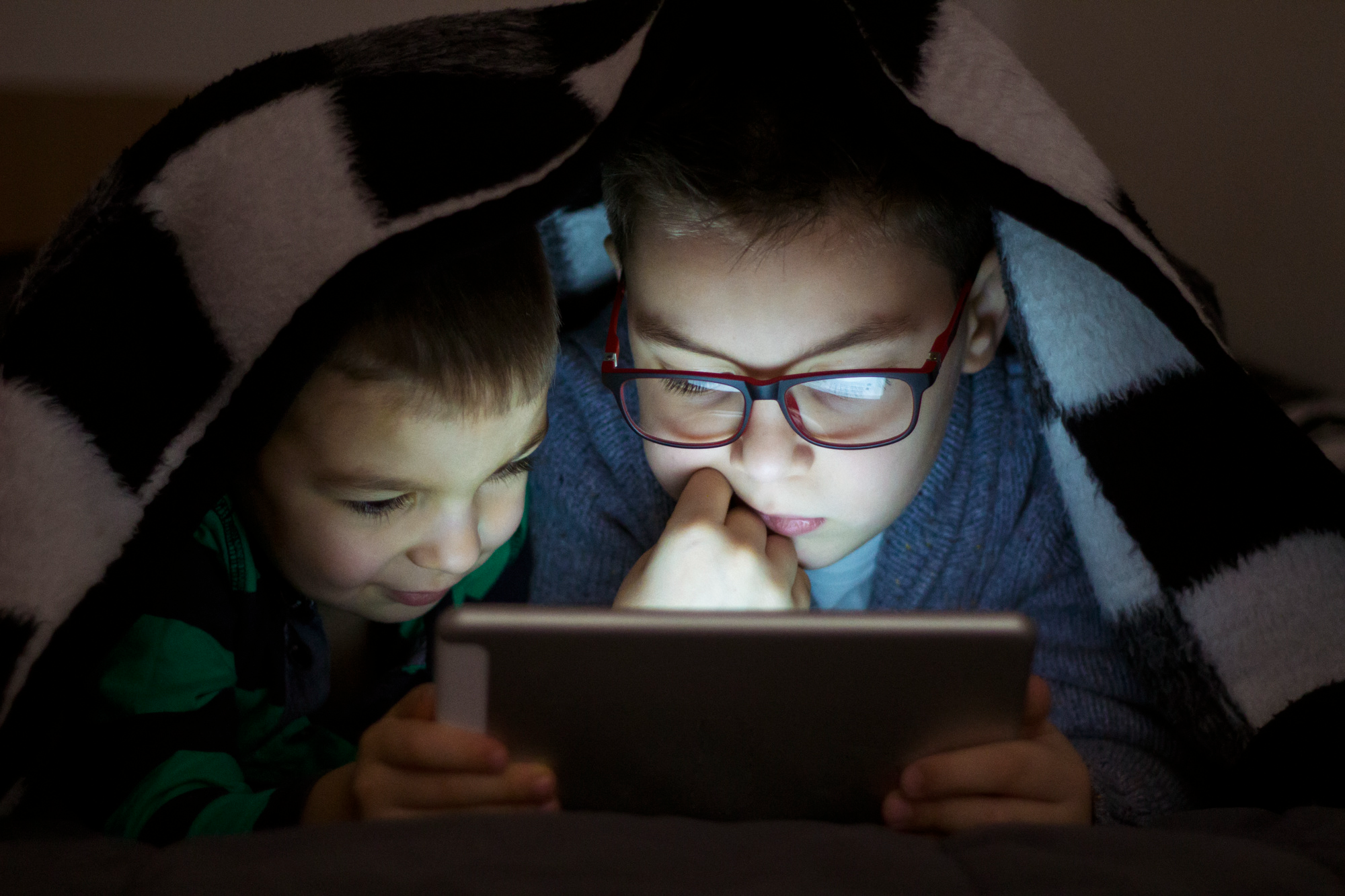 Two young kids sitting close together under a blanket in a dark room, watching something on a phone with the screen light shining on their faces.