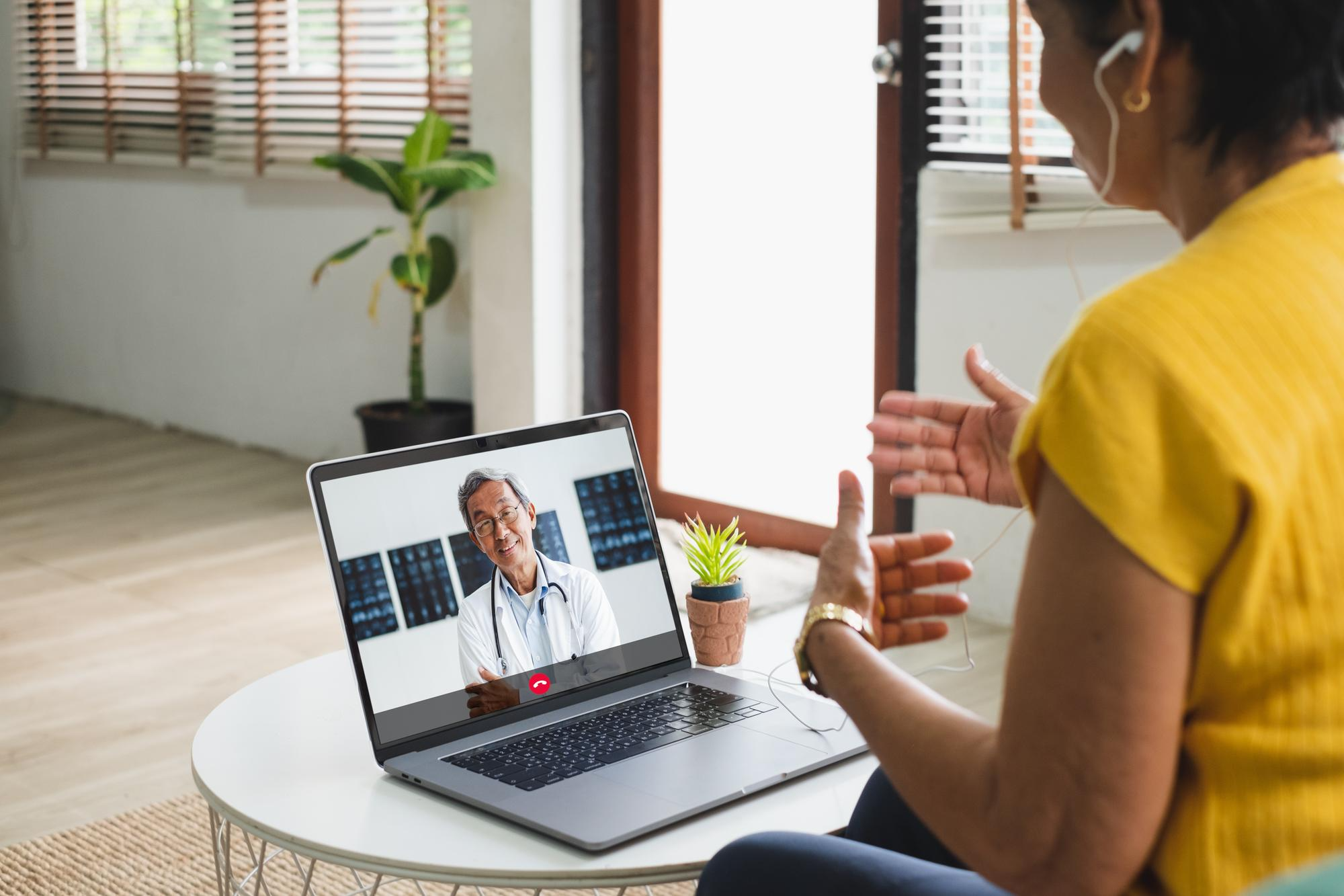 A woman sitting at home receiving online therapy, talking to a doctor visible on her laptop screen during a virtual counselling session.