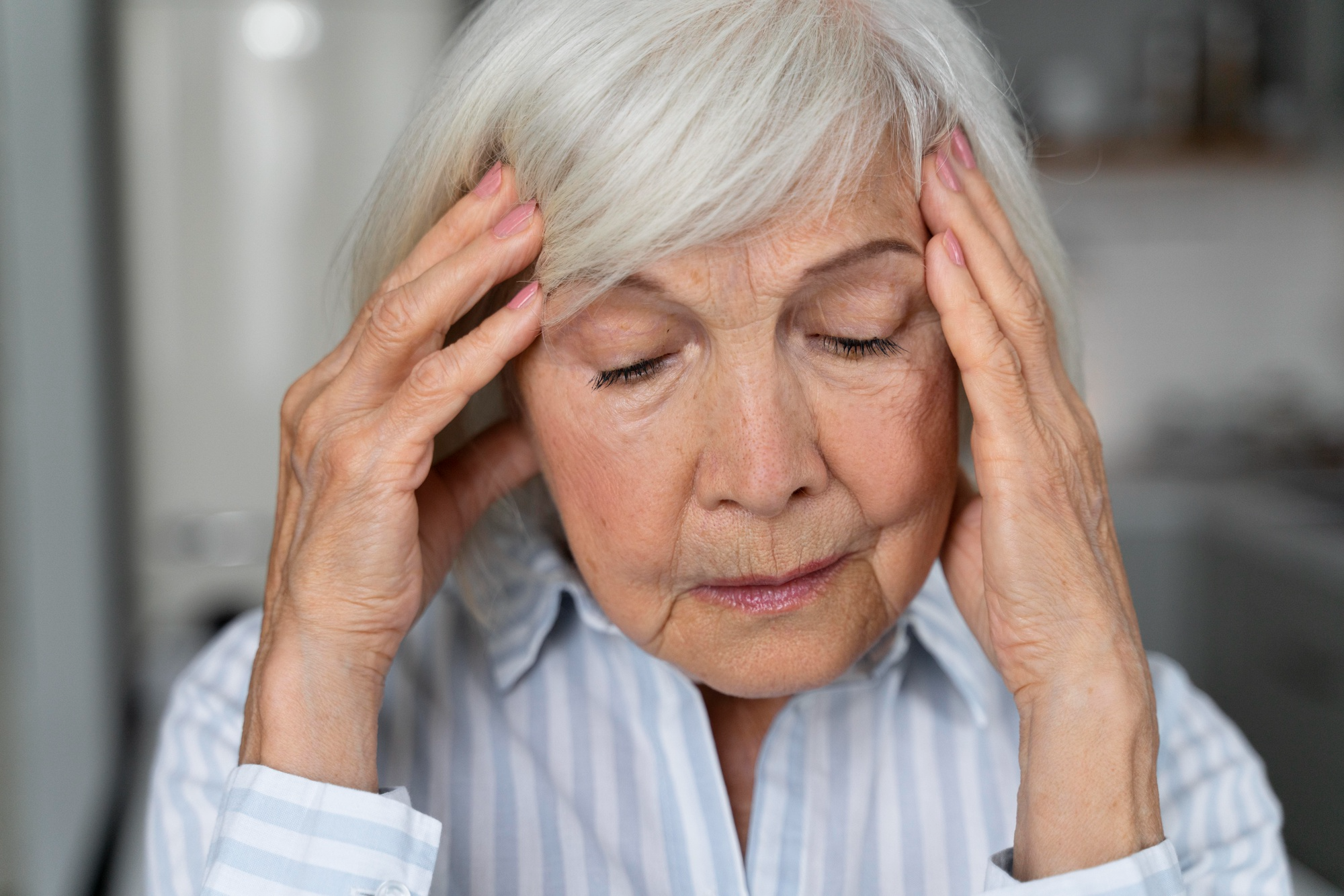 An old woman with grey hair who has Alzheimer’s Disease is holding her head as she is trying to remember something, looking troubled.