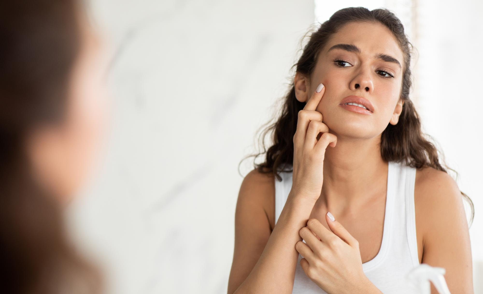 A woman closely looks at her reflection in the mirror, noticing uneven tone and fine lines on her face.