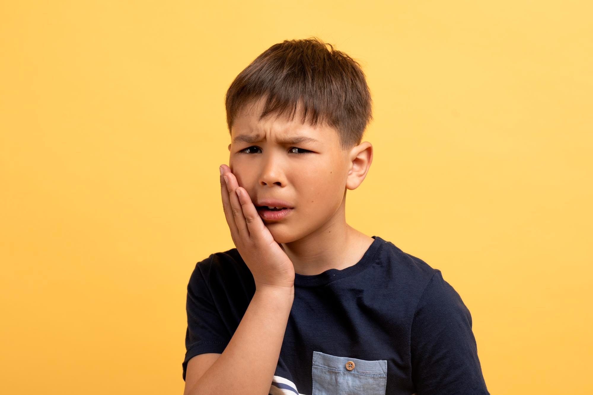 Young child touching their cheek in discomfort, showing signs of tooth pain caused by cavities, infection or poor dental hygiene.