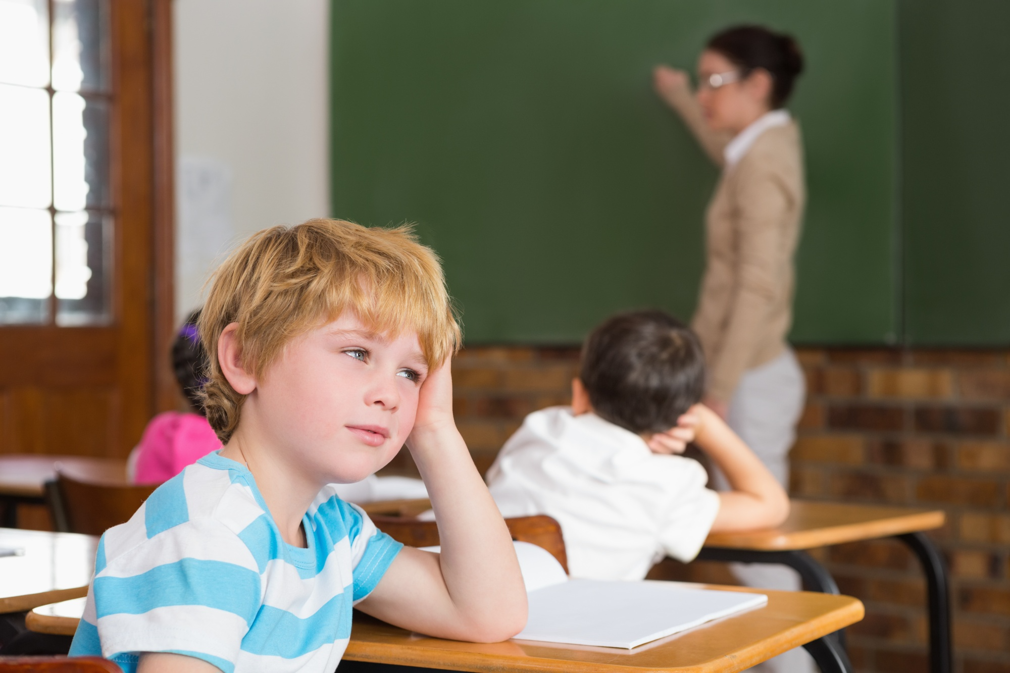 A school-aged child sits at a desk but gazes out the classroom window, appearing distracted and lost in thought while the teacher teaches.