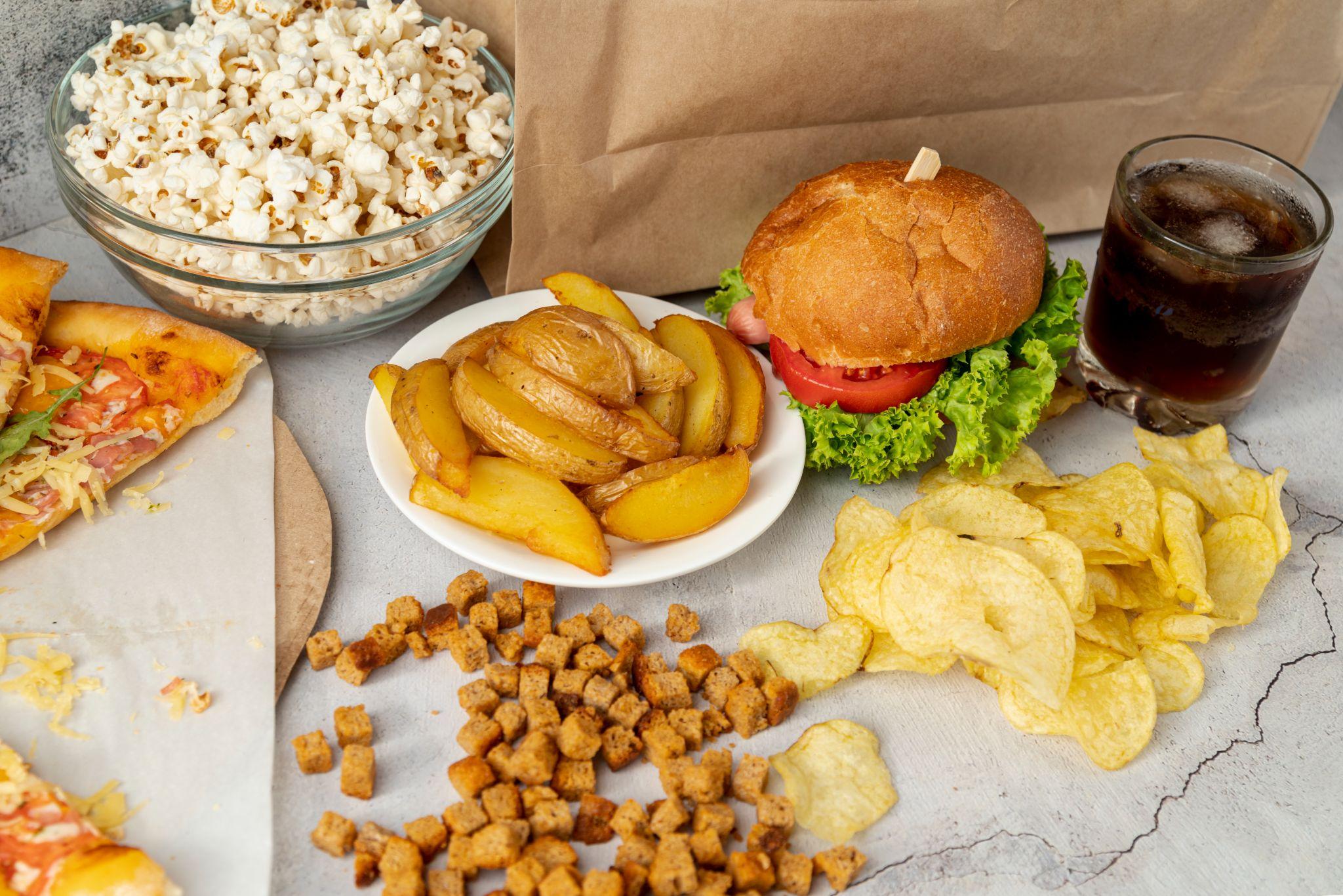 A top view of fast food items including burgers, fries, chips, popcorn, pizza, and soft drinks. Such oily and processed foods can trigger digestive problems and may worsen diarrhea symptoms.
