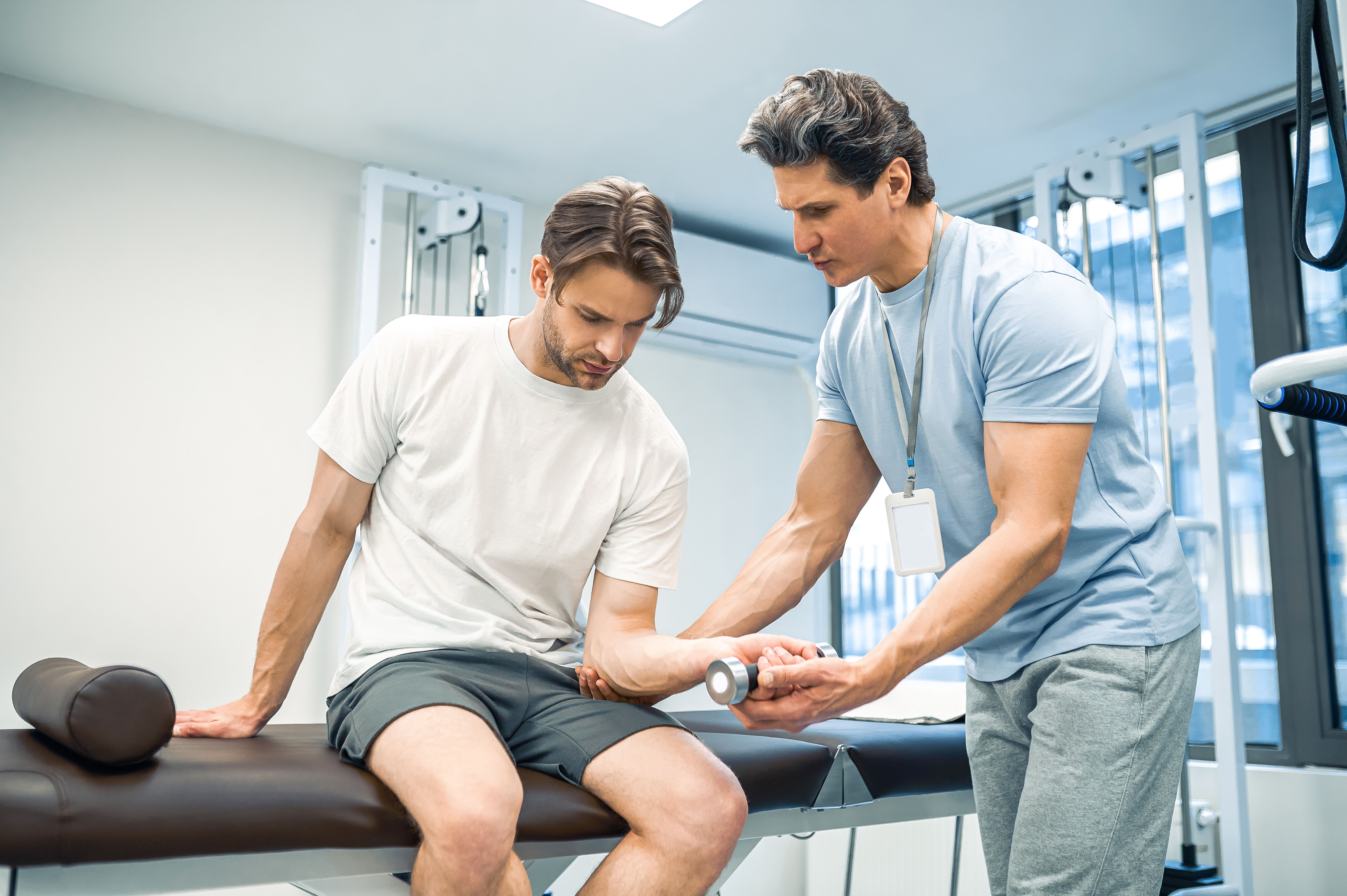 A physiotherapist assisting a male patient with arm rehabilitation using a light dumbbell in a clinical setting.