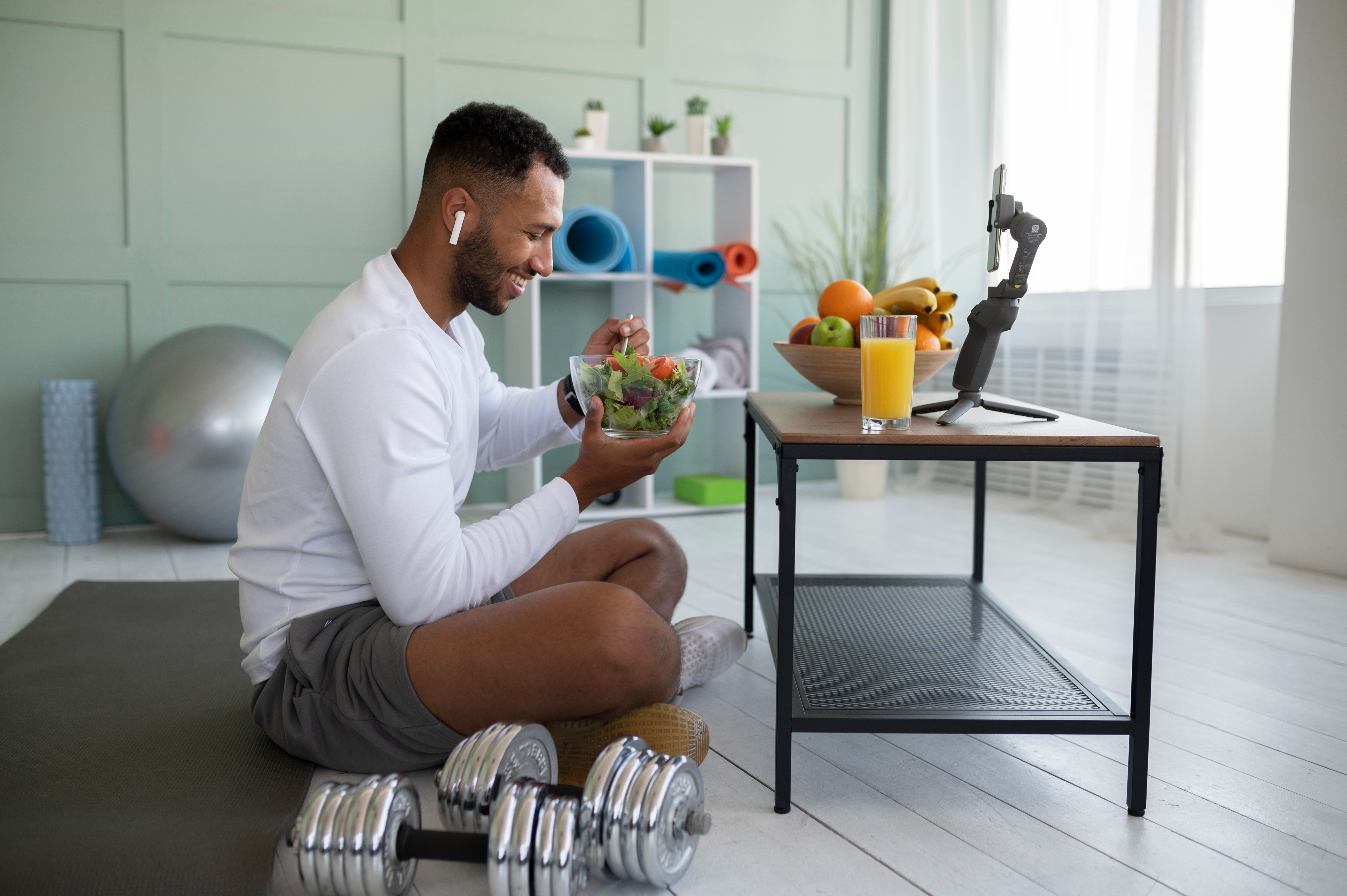 A young man enjoying a healthy salad and juice after a workout, seated on a yoga mat with dumbbells nearby.
