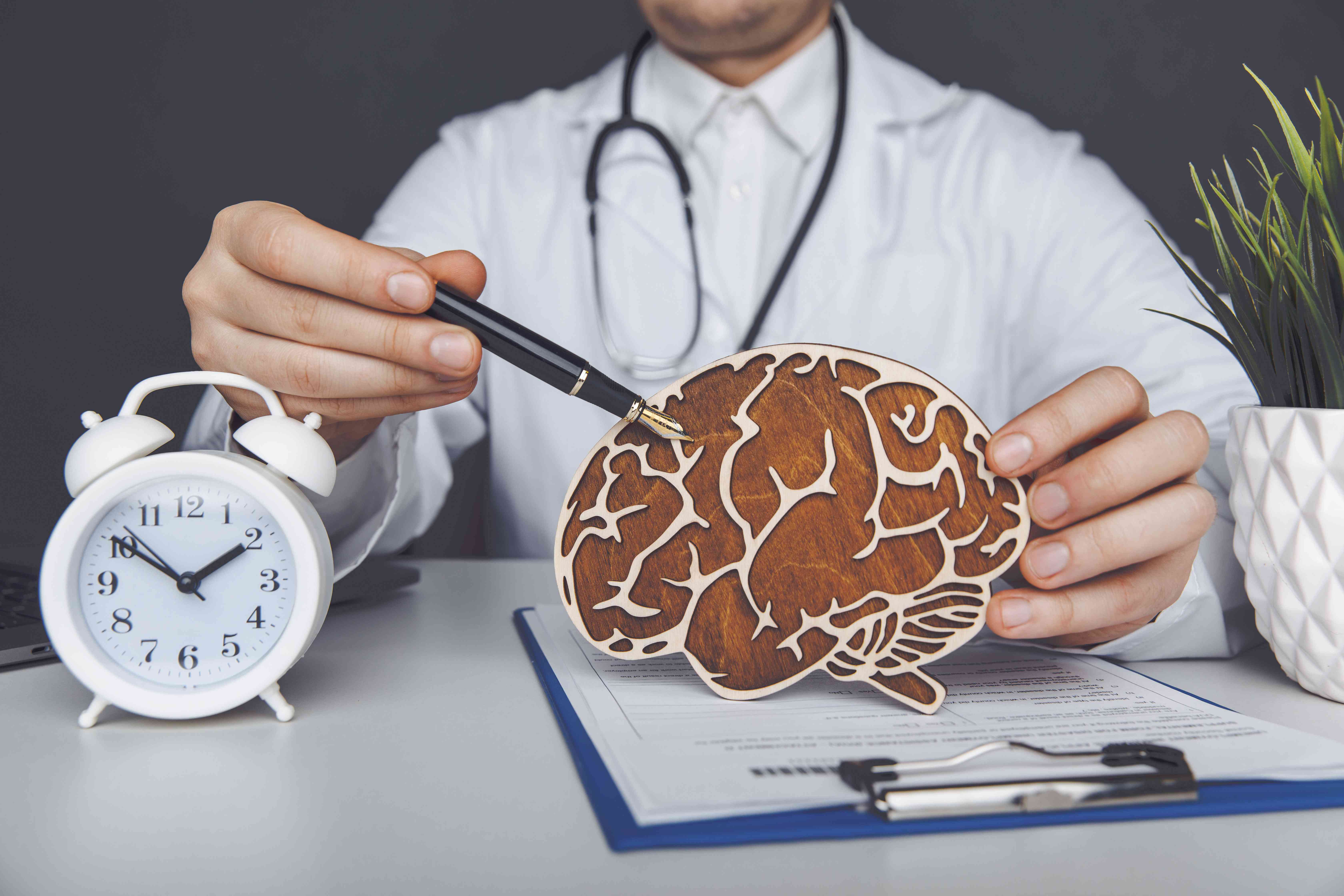 A doctor is pointing at a wooden brain model on a desk with a clipboard and clock, emphasizing brain health and neurology.