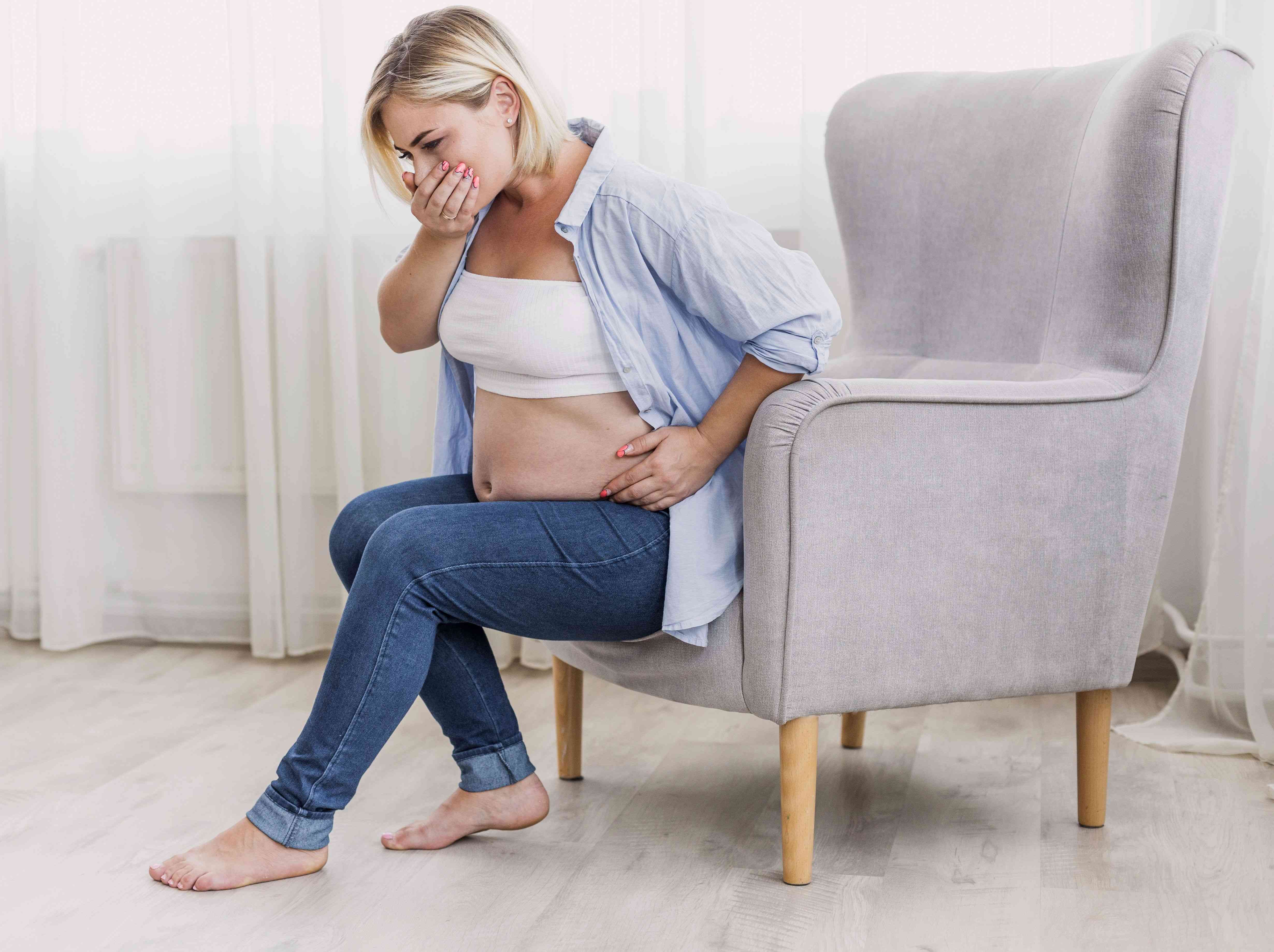 Pregnant woman sitting by a chair, holding her stomach and covering her mouth, experiencing nausea or abdominal discomfort.