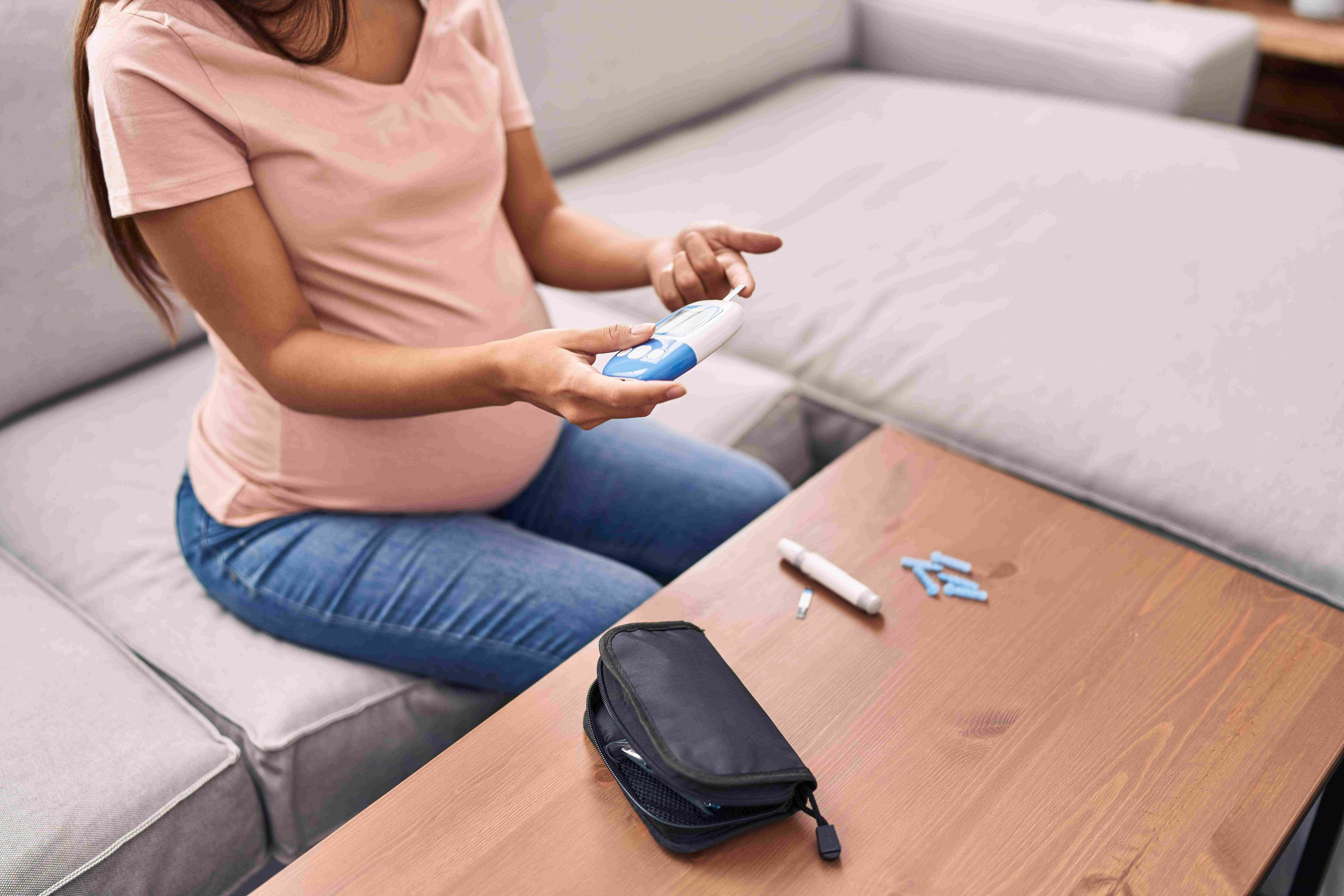 Pregnant woman sitting on a couch measuring her blood glucose levels using a glucometer.