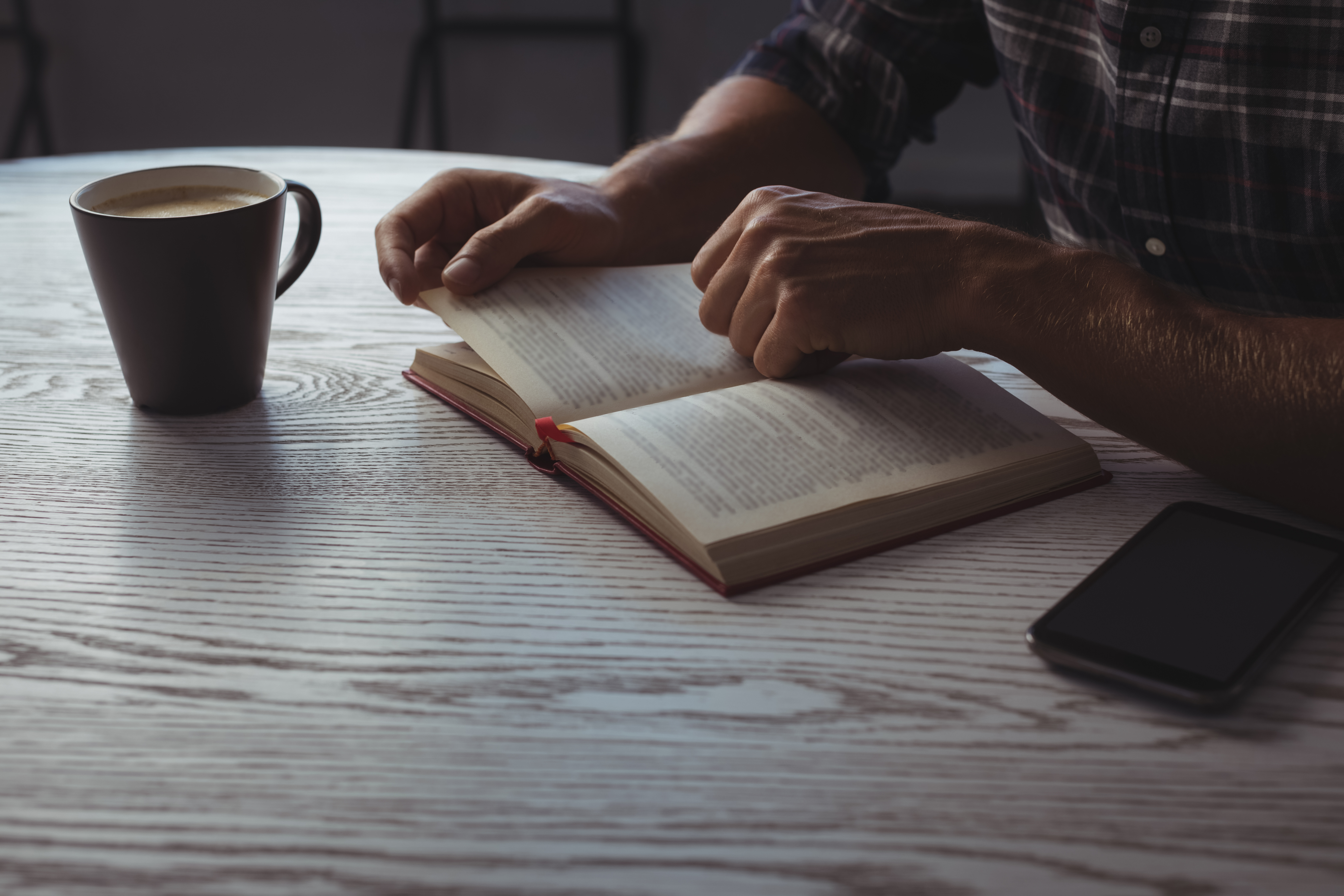 Man reading a book alone at a wooden table with coffee and a phone nearby.