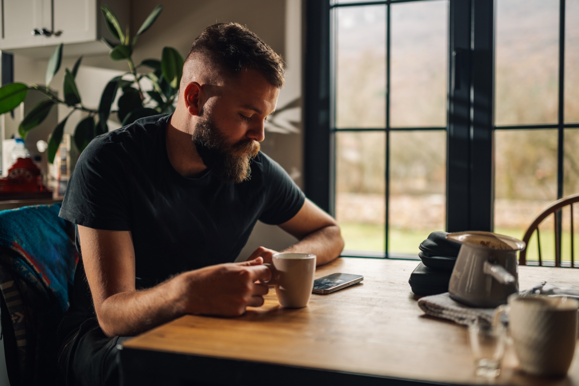 Bearded man in a dark t-shirt sitting at a wooden table, holding a coffee mug and looking thoughtful.