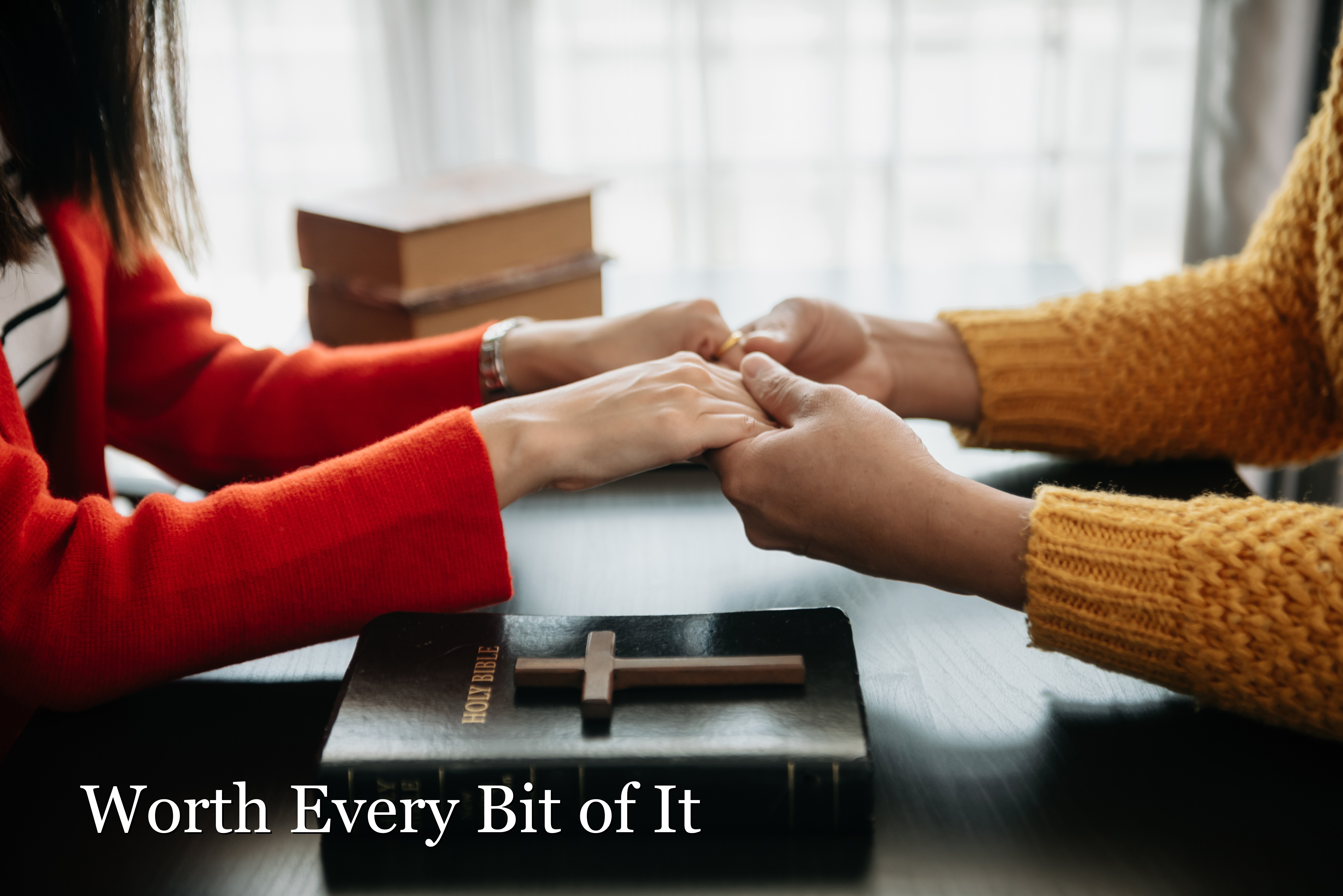 A married couple's clasped hands resting on an open Bible with a wooden cross, warm indoor light.