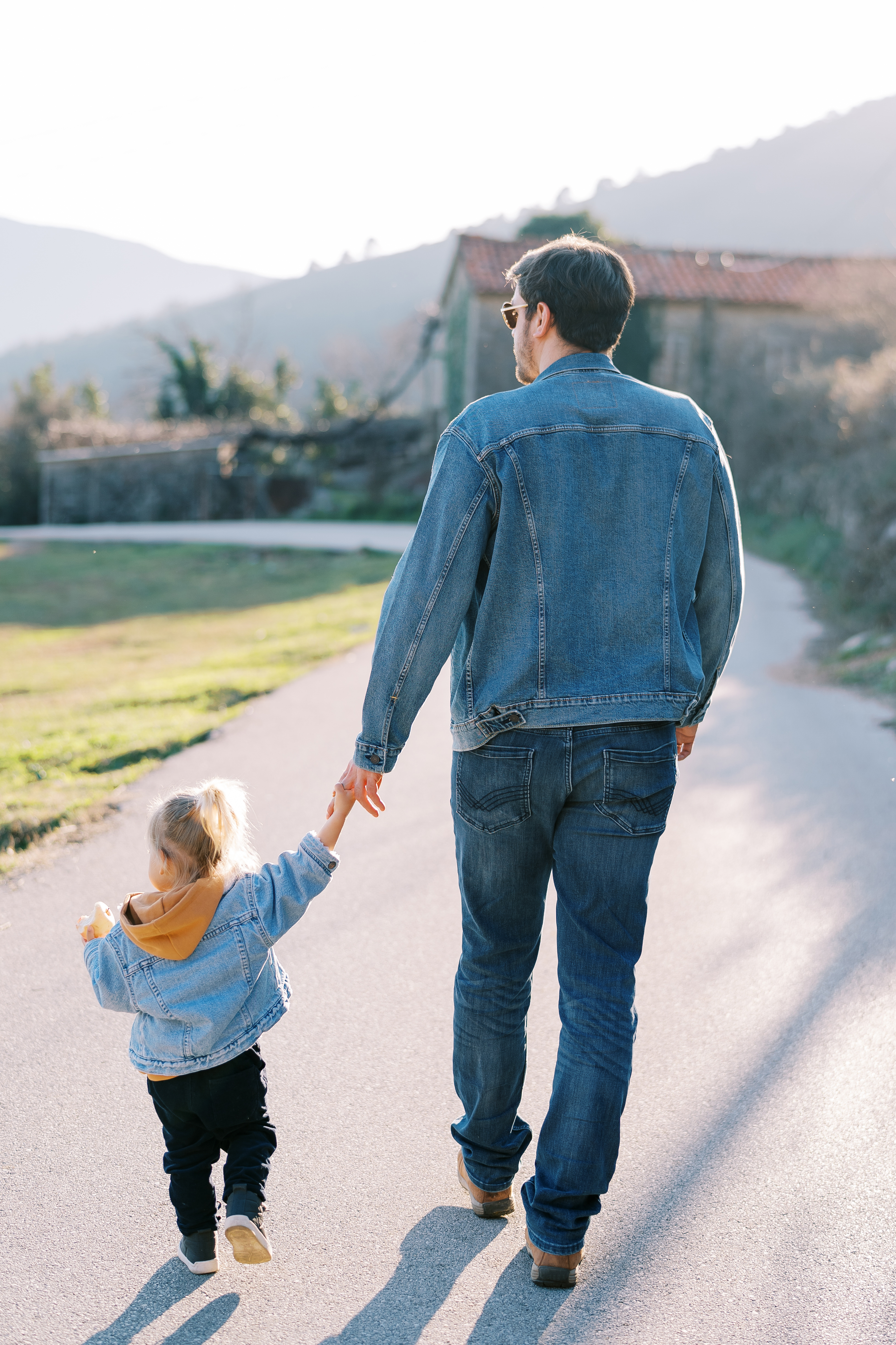 A father and young daughter walking hand in hand down a road, seen from behind