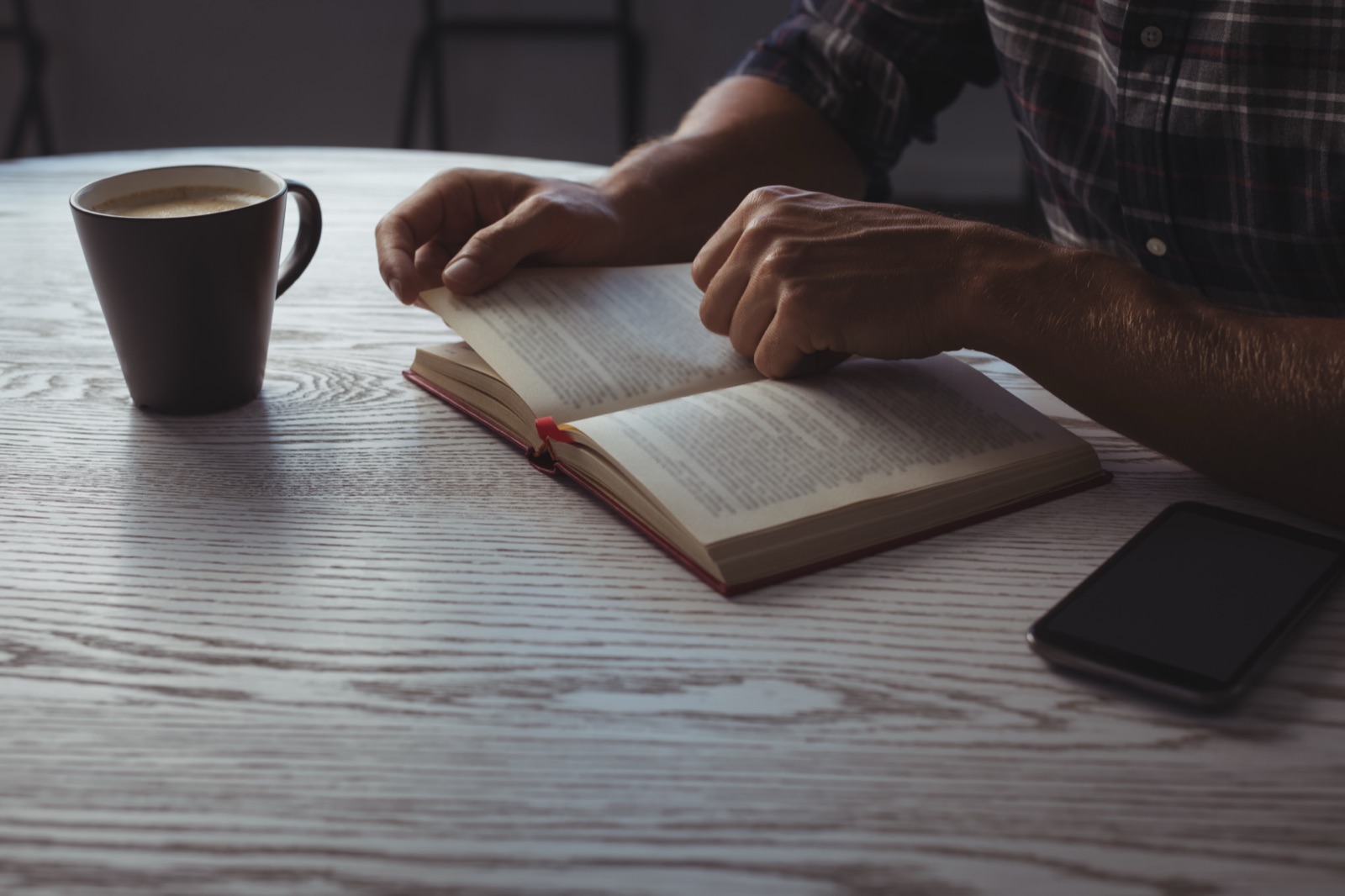 Man reading a book alone at a wooden table with coffee and a phone nearby.