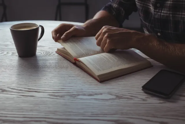 Man reading a book alone at a wooden table with coffee and a phone nearby.