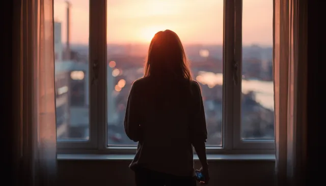 Silhouette of a woman gazing out a high-rise window at a glowing city sunset while holding her phone.