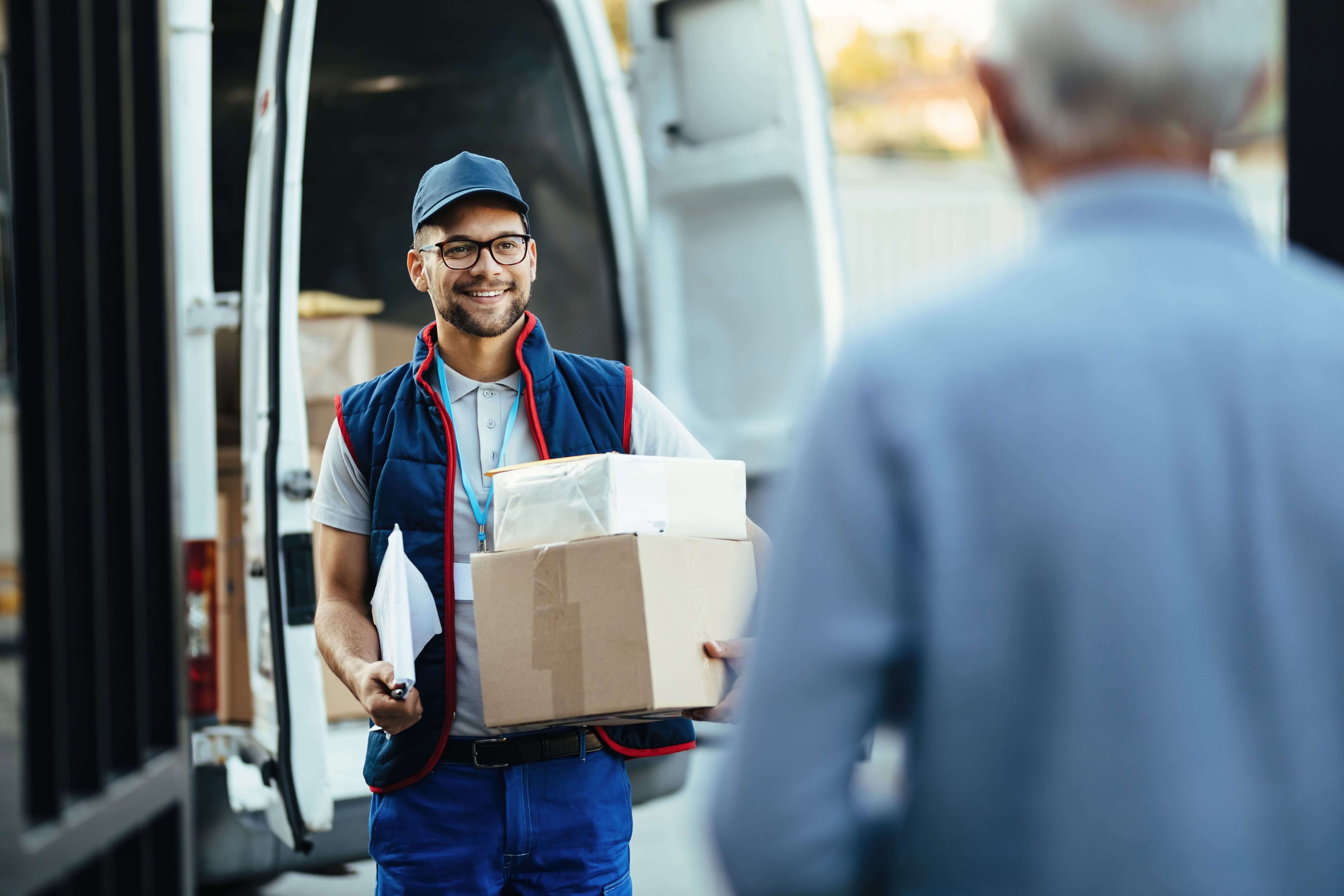 Happy delivery driver wearing uniform holding packages walking towards a man