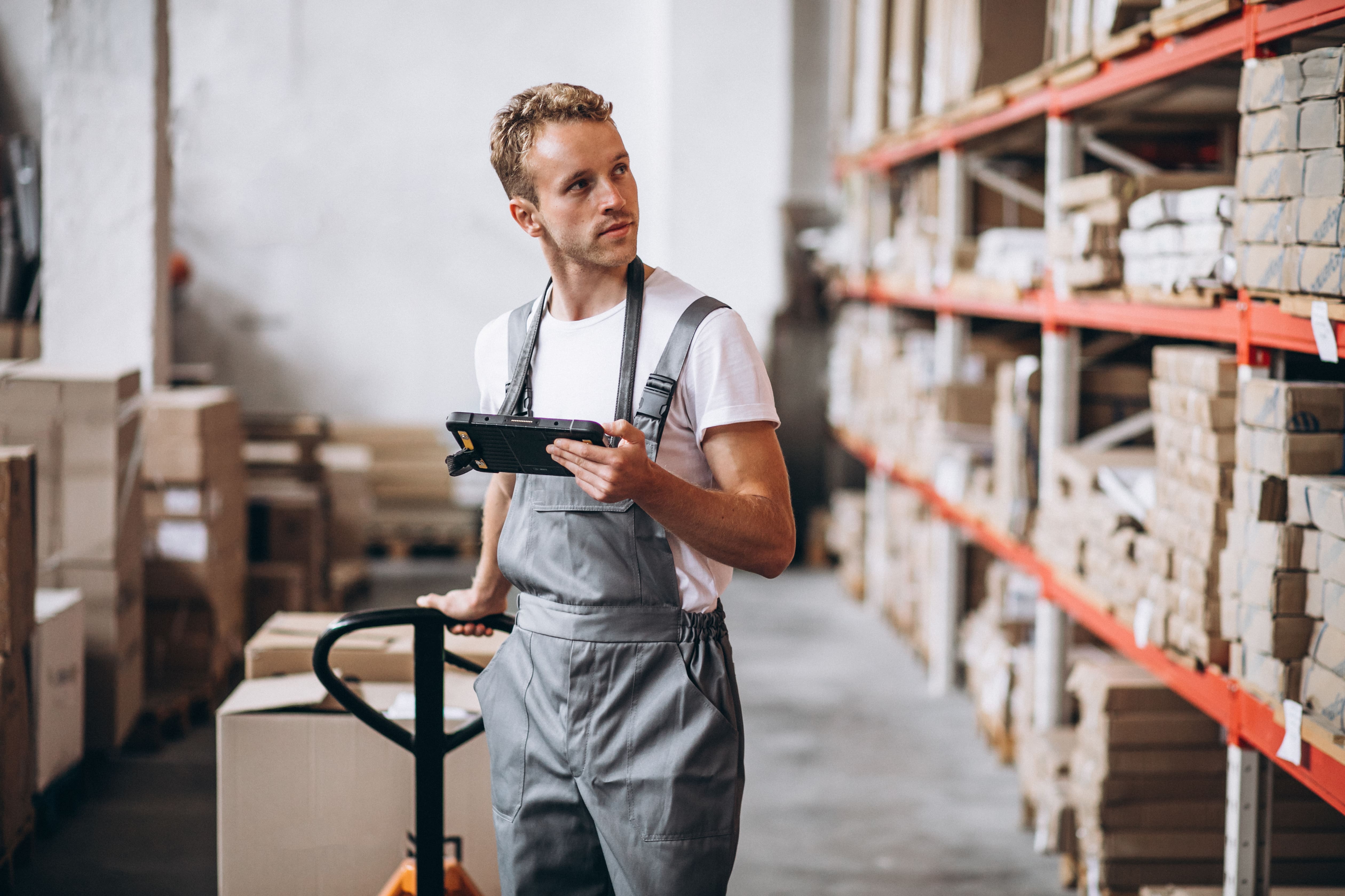 Man in warehouse pulling forklift behind him with a tablet in his hand, looking at the shelves