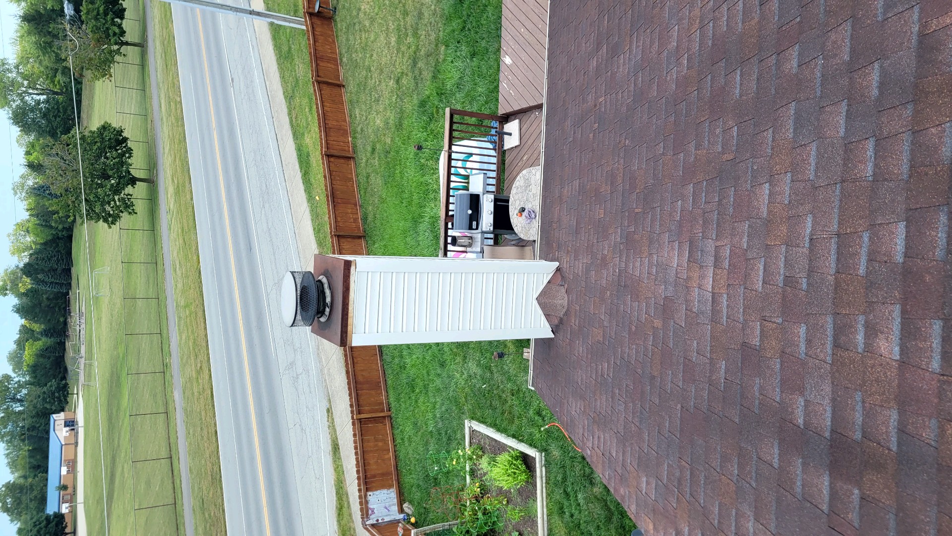 Rooftop view showing brown shingles, a white chimney with a metal cap, and surrounding backyard with outdoor furniture