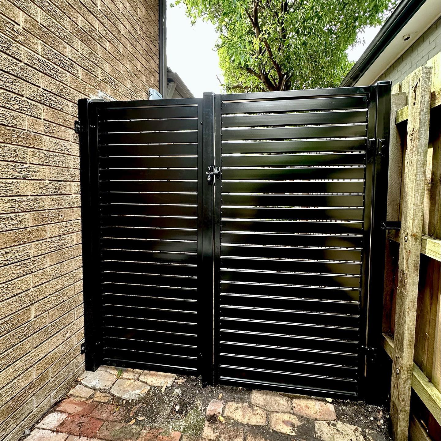 Two black slat steel gates between brick wall and wooden fence