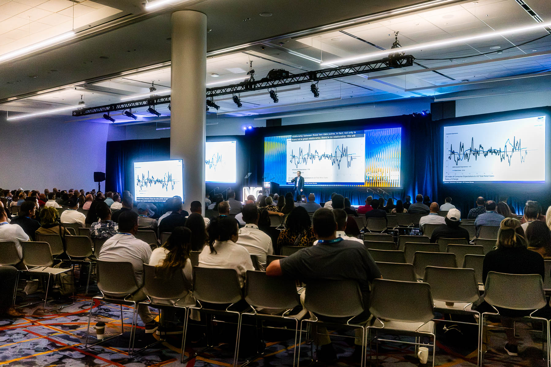A speaker presents on a large stage with multiple screens displaying graphs to a seated audience in a conference hall.
